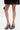 A person wearing a short black lace skirt stands on a white background, holding a small black handbag and sporting AKIRA LAMONT SNAKE SANDAL CHUNKY PLATFORM heels.