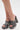 A close-up of a person wearing the AKIRA LAMONT SNAKE SANDAL CHUNKY PLATFORM, featuring gray faux leather with a snakeskin pattern and open toes, standing on a white surface.