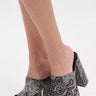 A close-up of a person wearing the AKIRA LAMONT SNAKE SANDAL CHUNKY PLATFORM, featuring gray faux leather with a snakeskin pattern and open toes, standing on a white surface.