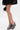 A person in a black lace dress stands with legs crossed, wearing AKIRA LAMONT SNAKE SANDAL CHUNKY PLATFORM gray sandals and holding a small black handbag with keychain accents against a plain white background.
