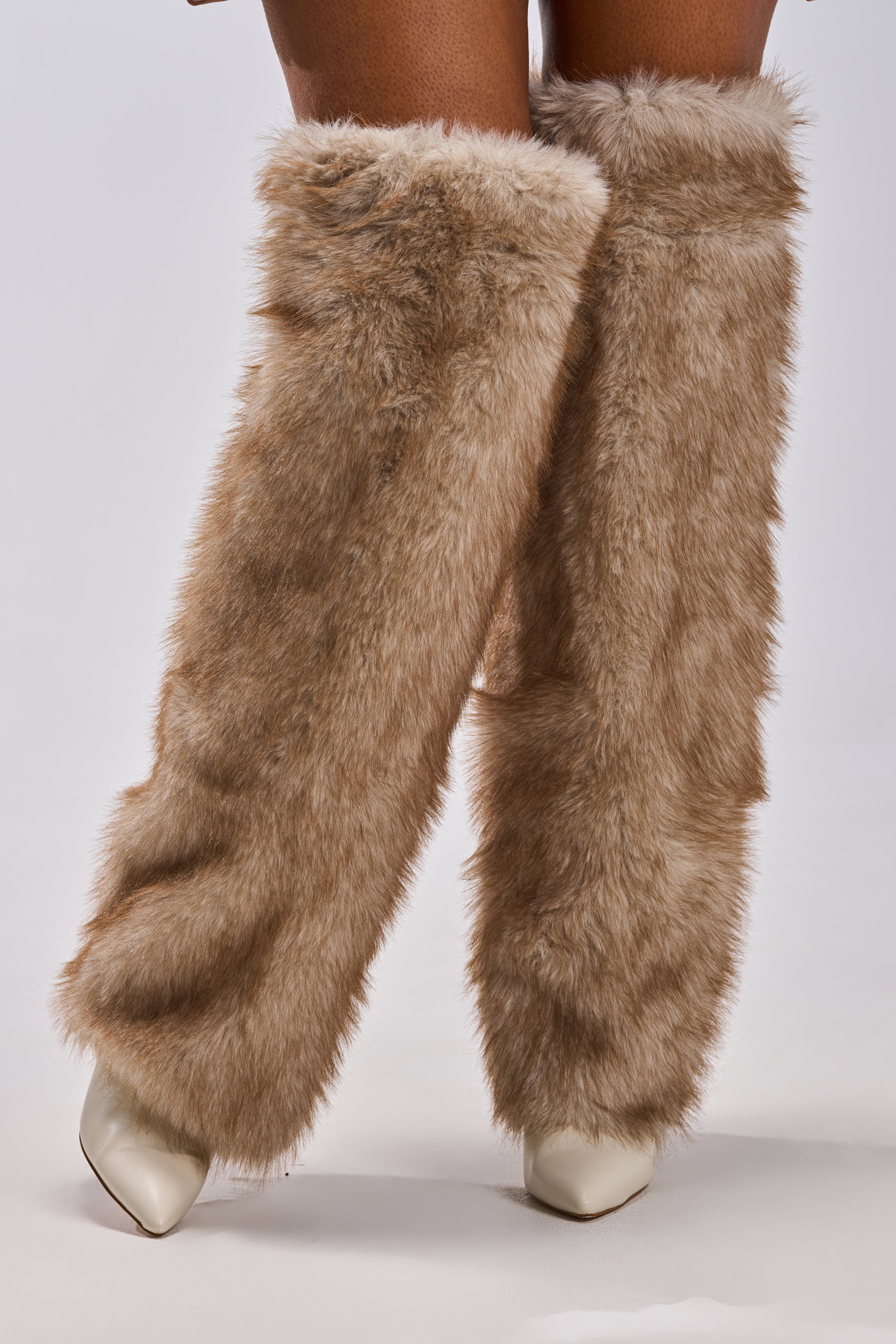 A person wears the AZALEA WANG LAGANN BONE THIGH HIGH FUR LUXE BOOT—tall, fluffy light brown faux fur boots over cream stiletto heels—standing against a plain light background.