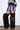 A person in a light purple faux fur mini skirt, matching bandeau, sheer black long-sleeve underlayer, and AZALEA WANG LANGLEYE BLACK THIGH HIGH FUR LUXE BOOTS stands against a plain white wall.