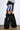 A person in a fuzzy grey crop top, lavender faux fur mini skirt, black mesh sleeves, and AZALEA WANG LANGLEYE BLACK THIGH HIGH FUR LUXE BOOTS stands by a white wall and brown floor.
