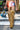 A person in a colorful mini skirt and AZALEA WANG LANGDON BROWN THIGH HIGH FUR LUXE BOOTS with stiletto heels walks across a city street, carrying a shiny blue handbag. Urban buildings and a 