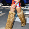 Someone walks down a city street in the AZALEA WANG LANGDON BROWN THIGH HIGH FUR LUXE BOOT, paired with orange and blue shorts and a white crop top, with a blue SUV visible in the background.