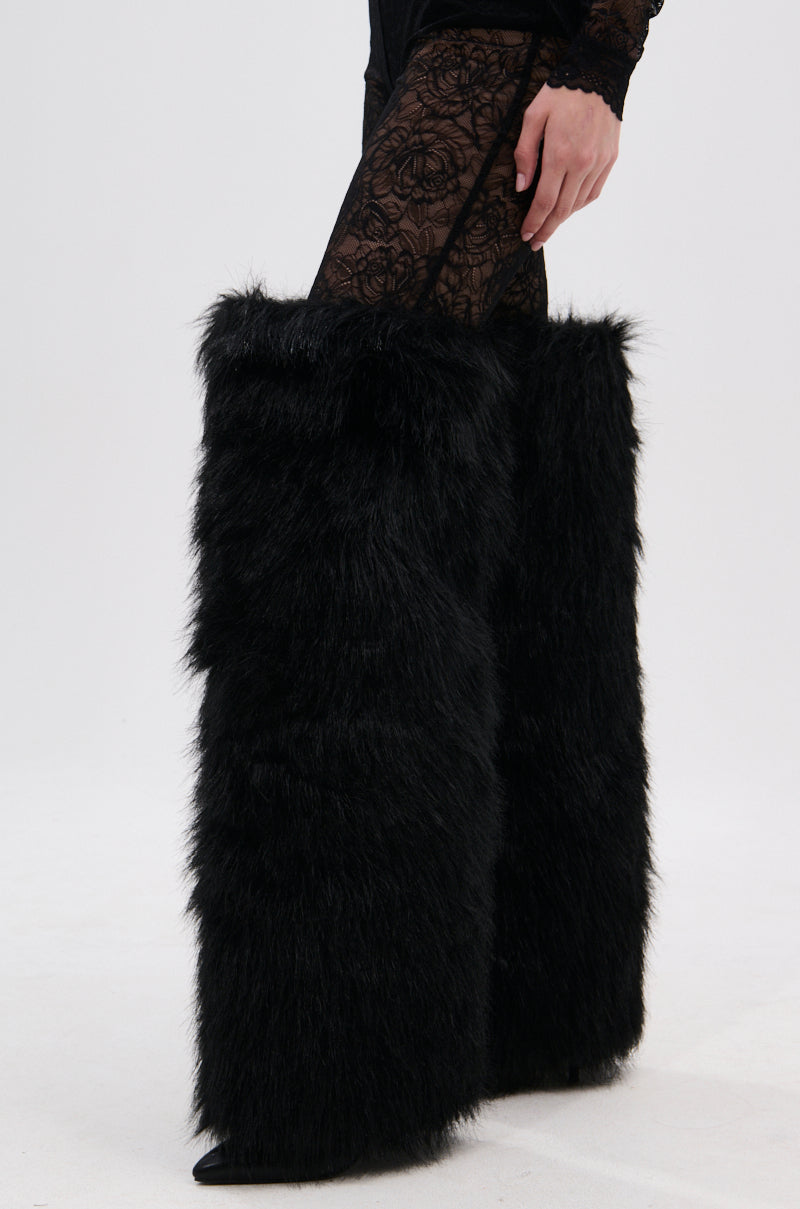 A person stands on a white surface wearing the AZALEA WANG LANGLEYE BLACK THIGH-HIGH FUR LUXE BOOT, styled with black lace-patterned leggings and oversized faux fur leg warmers from thigh to ankle.