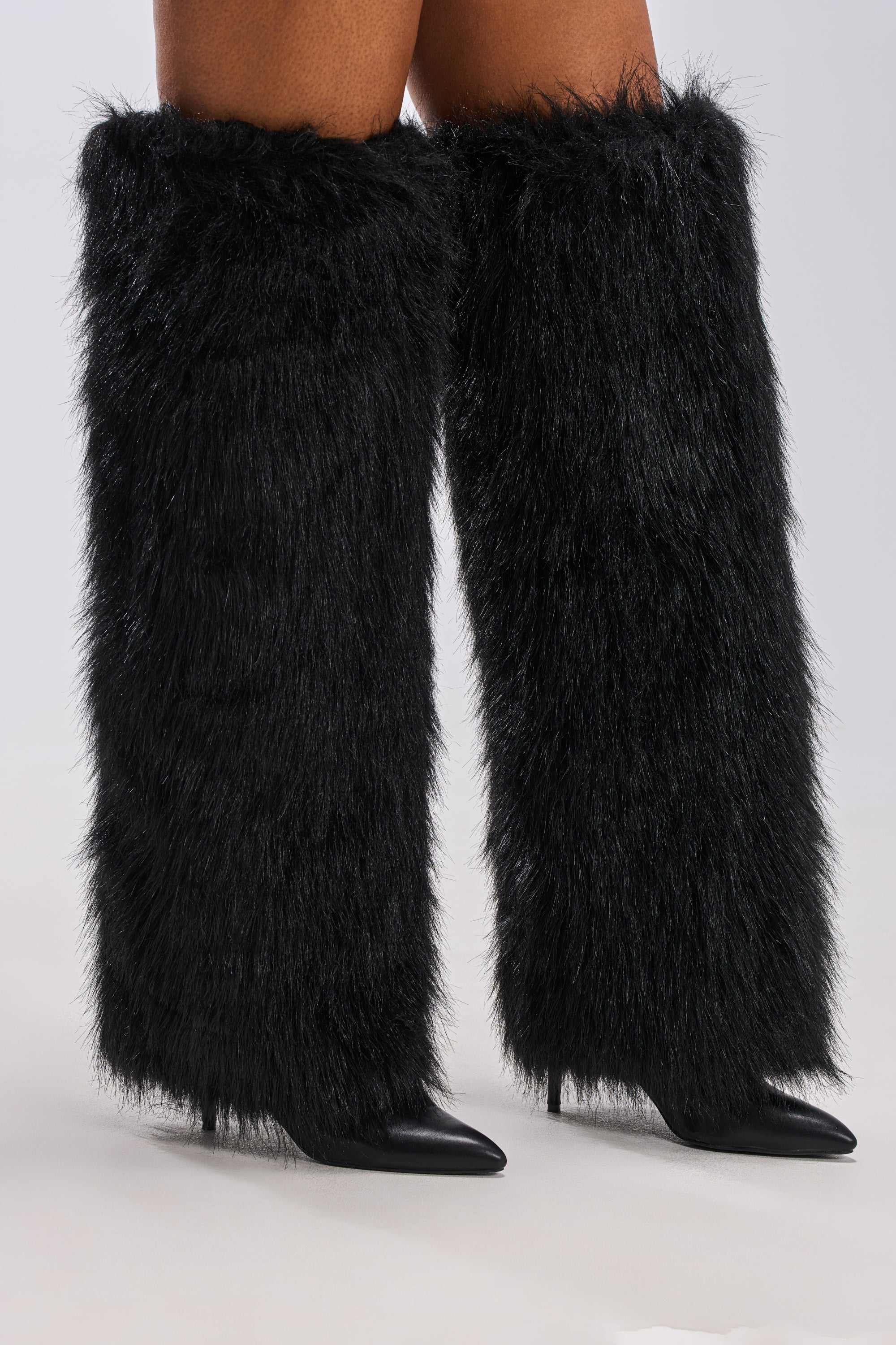 A pair of legs wearing AZALEA WANG LANGLEYE Black Thigh High Fur Luxe Boots, featuring faux fur, pointed toes, and stiletto heels against a plain light background.