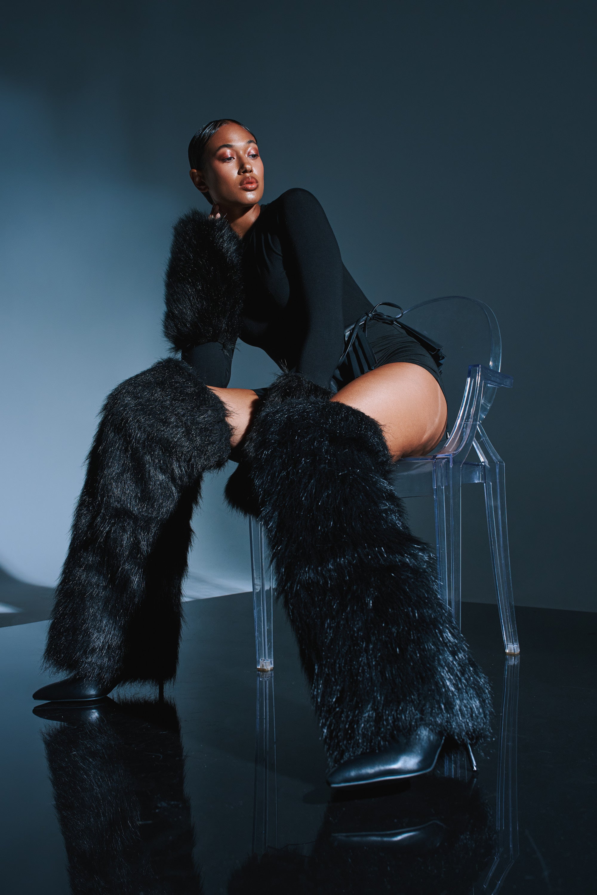 A confident woman in a black outfit and AZALEA WANG LANGLEYE BLACK THIGH HIGH FUR LUXE BOOTS sits on a clear acrylic chair against a dark background, gazing upward with a poised, stylish expression.