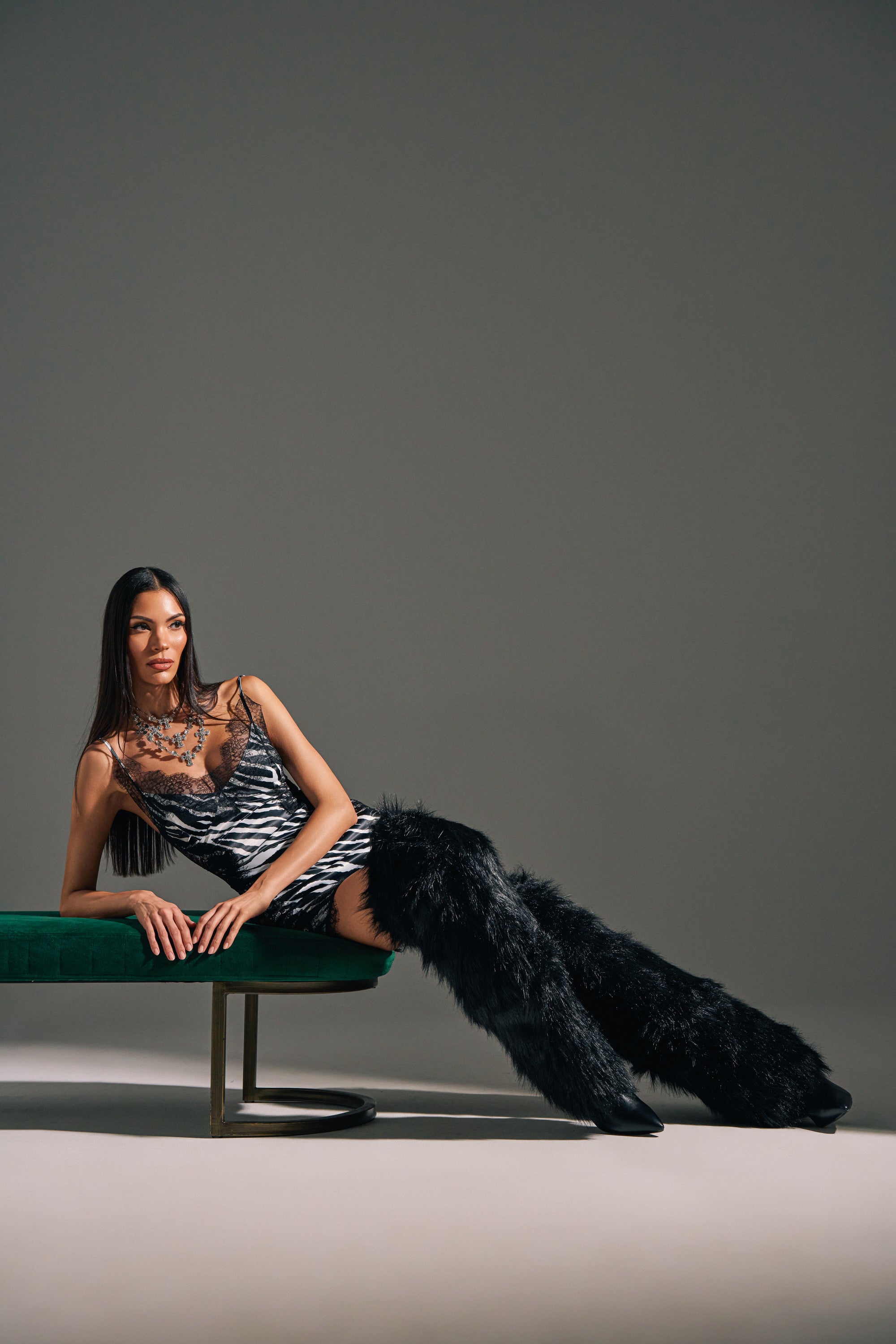 A woman with long dark hair lounges on a green bench, wearing a zebra-striped mini dress and striking AZALEA WANG LANGLEYE BLACK THIGH HIGH FUR LUXE BOOTS. The background is plain gray.