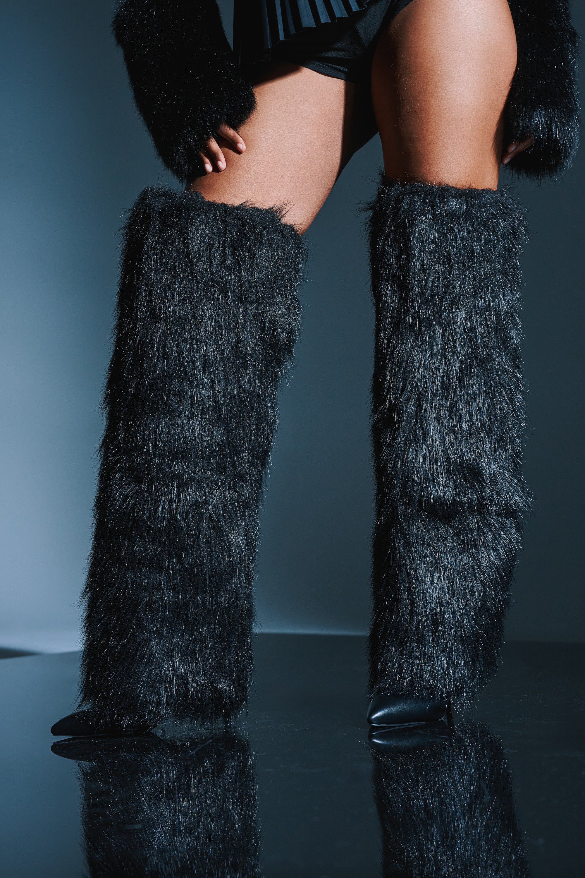 A person models the AZALEA WANG LANGLEYE BLACK THIGH HIGH FUR LUXE BOOT, standing on a reflective surface with their legs and lower body shown against a gray background.