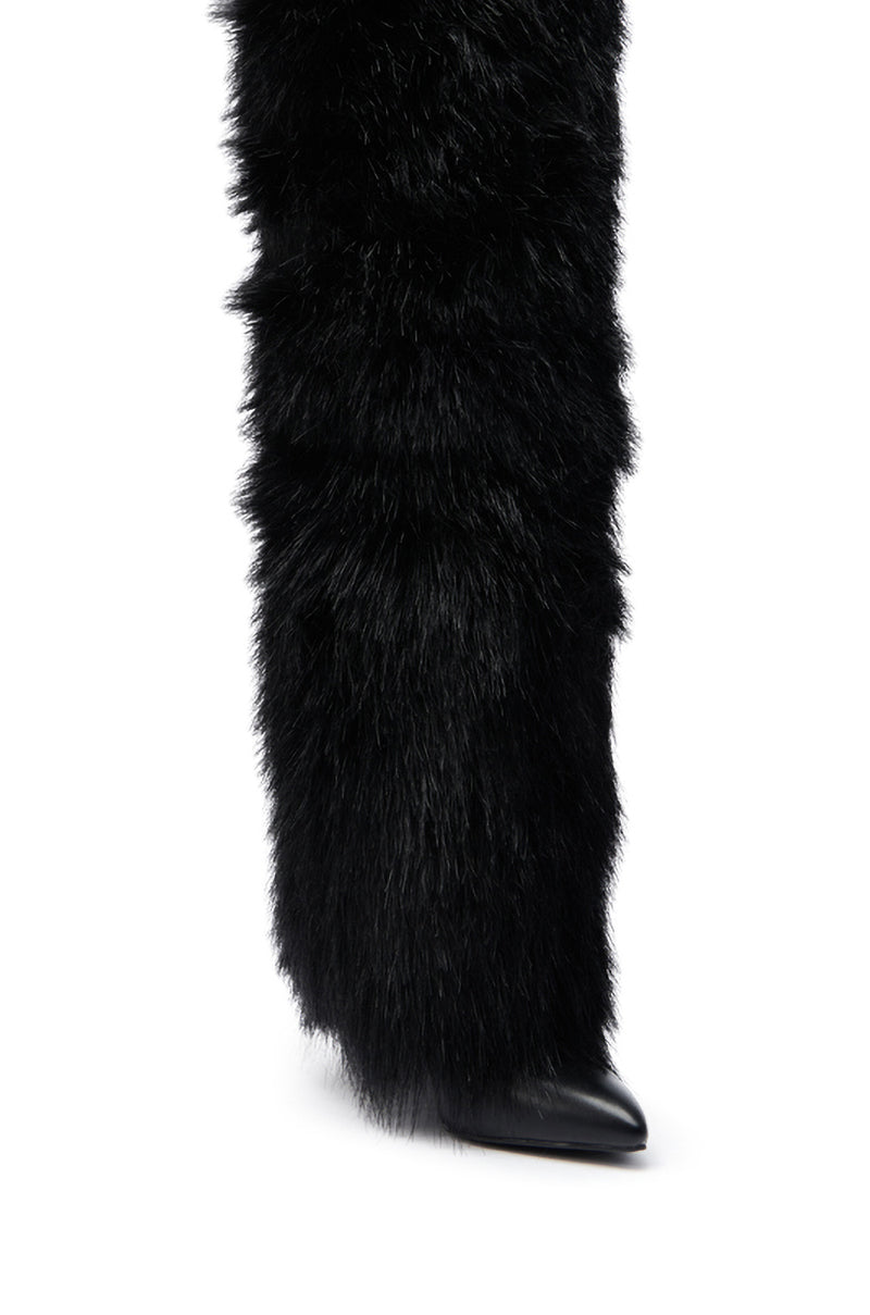 A close-up of a person's lower leg shows the AZALEA WANG LANGLEYE BLACK THIGH-HIGH FUR LUXE BOOT, featuring thick, long black faux fur that covers most of a pointed shoe for a bold textured style.