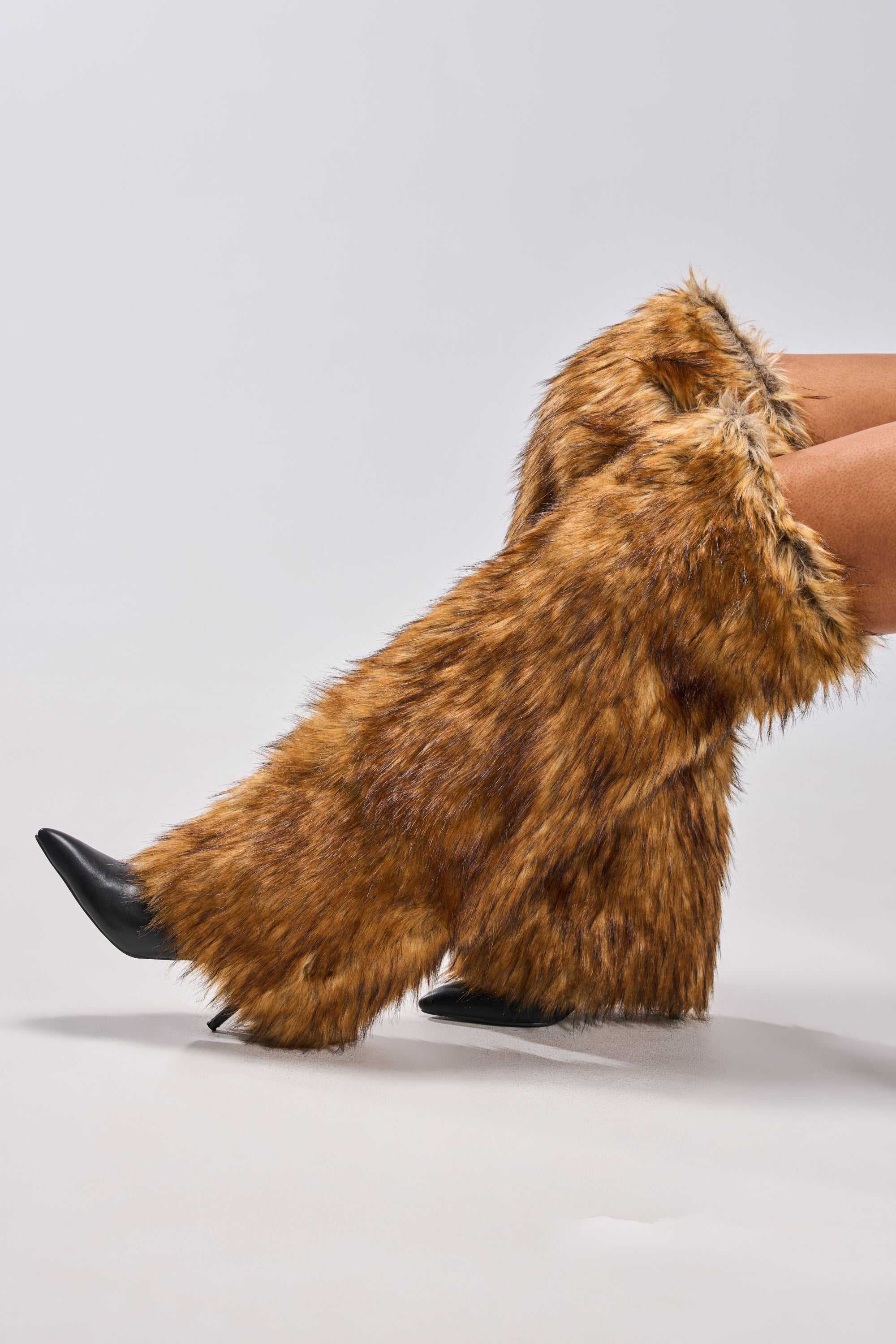 A person wearing AZALEA WANG LANGLEYE BROWN THIGH HIGH FUR LUXE BOOT stilettos with tall, shaggy brown faux fur, styled over black boots against a plain light gray background.