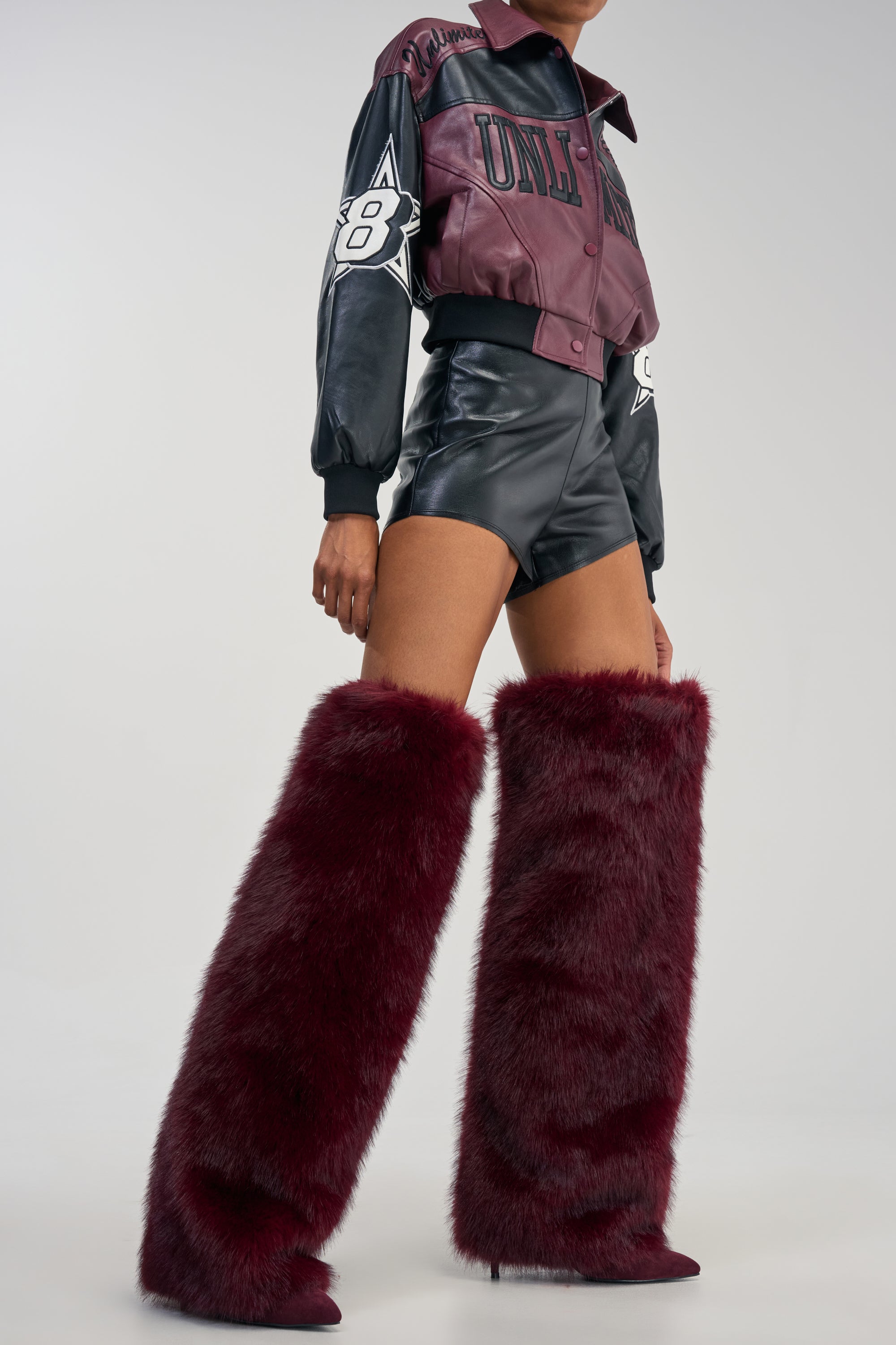 A person wears black leather shorts, a maroon and black jacket, and the AZALEA WANG LANGLEYE BURGUNDY FUR THIGH HIGH BOOTS. The background is plain and light grey.