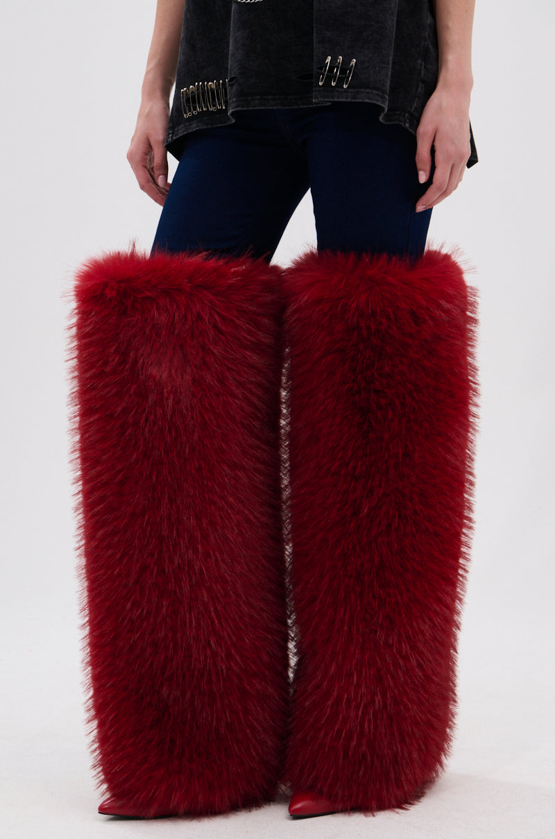 A person wearing the AZALEA WANG LANGLEYE RED THIGH-HIGH FUR LUXE BOOT—tall, fluffy red stiletto boots reaching above the knees—paired with dark pants and a black top. Only the lower half of their body is shown.