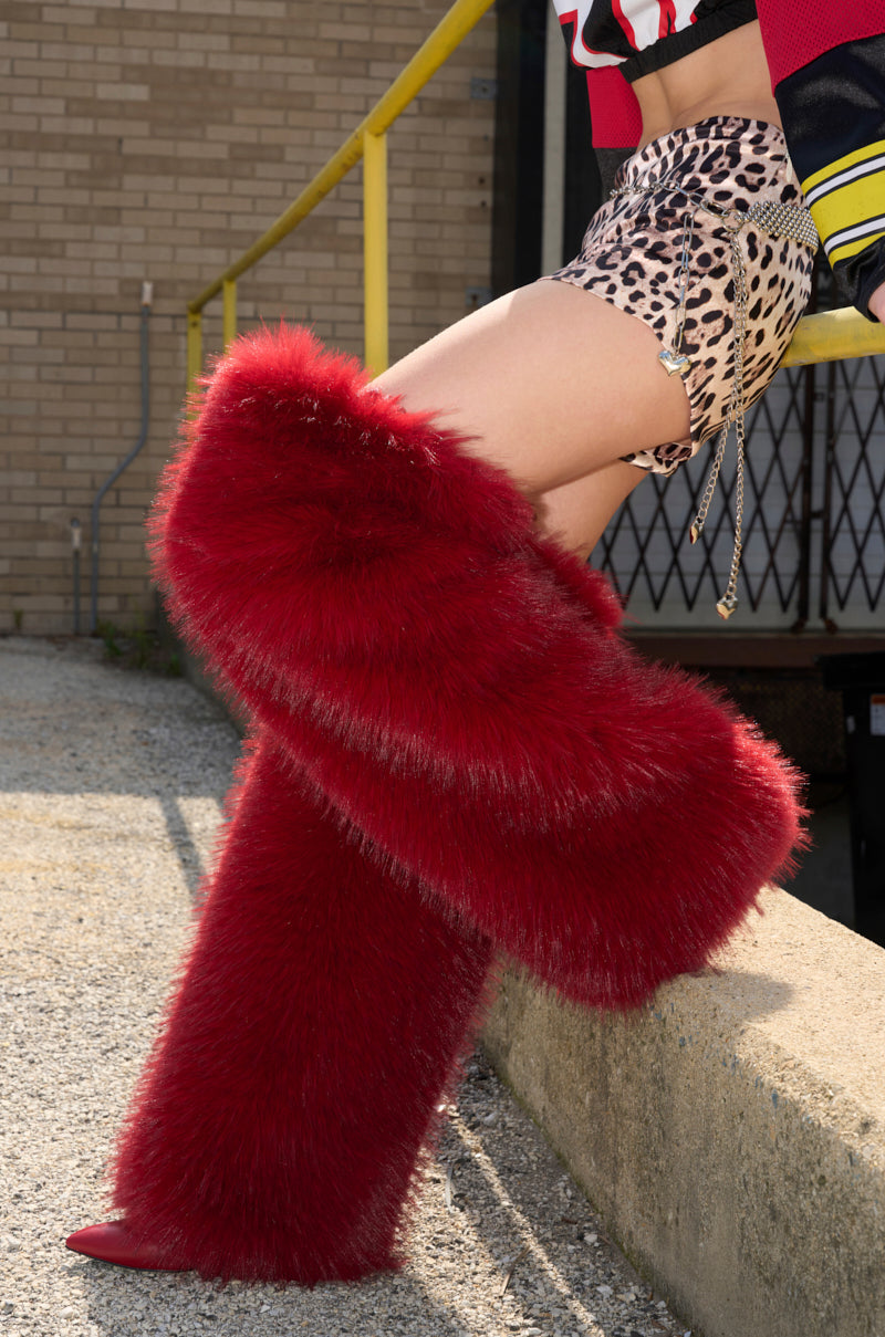 Wearing AZALEA WANG LANGLEYE RED THIGH HIGH FUR LUXE BOOTS and leopard print shorts, a person stands with one leg crossed on a concrete ledge near a yellow rail and brick building.
