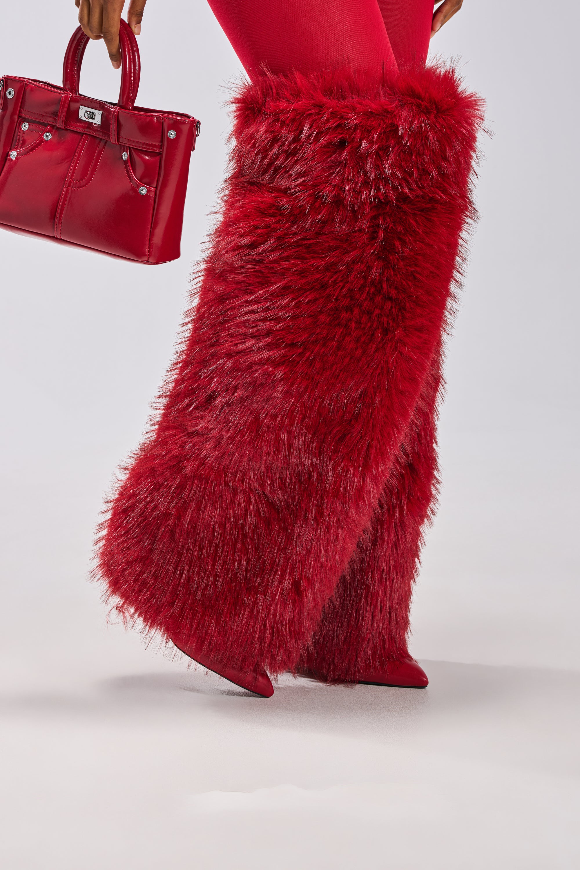 A person wears the AZALEA WANG LANGLEYE RED THIGH HIGH FUR LUXE BOOT with red leggings and holds a shiny red handbag, making the bold boots and matching bag pop against a plain, light background.