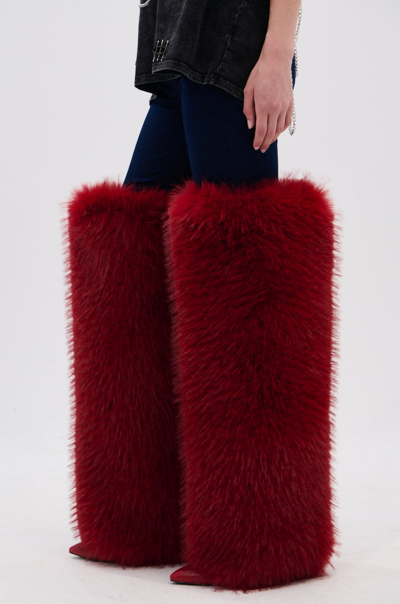 A person is seen from the waist down wearing the AZALEA WANG LANGLEYE RED THIGH-HIGH FUR LUXE BOOT—oversized, fluffy red faux fur boots that cover their legs from knees to feet, paired with dark pants and a black top.
