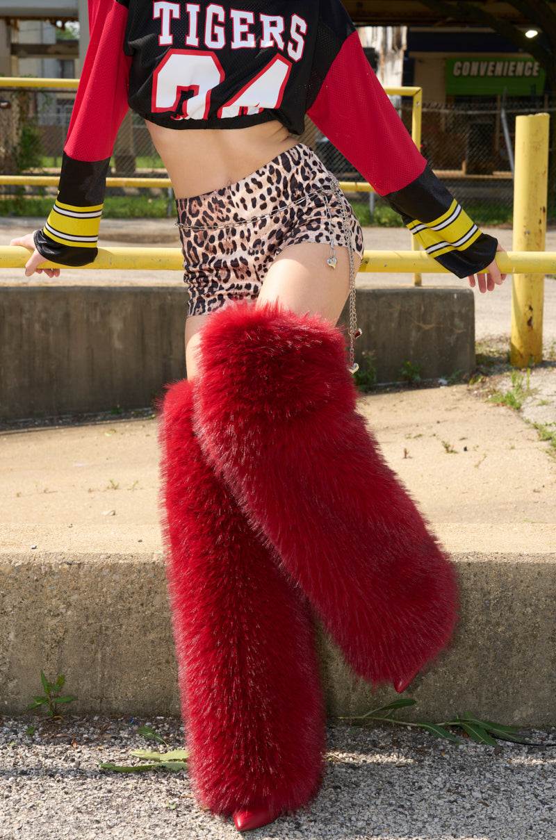 A person poses outdoors in a cropped "Tigers 84" jersey, leopard print shorts, and AZALEA WANG LANGLEYE RED THIGH HIGH FUR LUXE BOOTS, standing on one leg with the other slightly raised near a concrete curb.