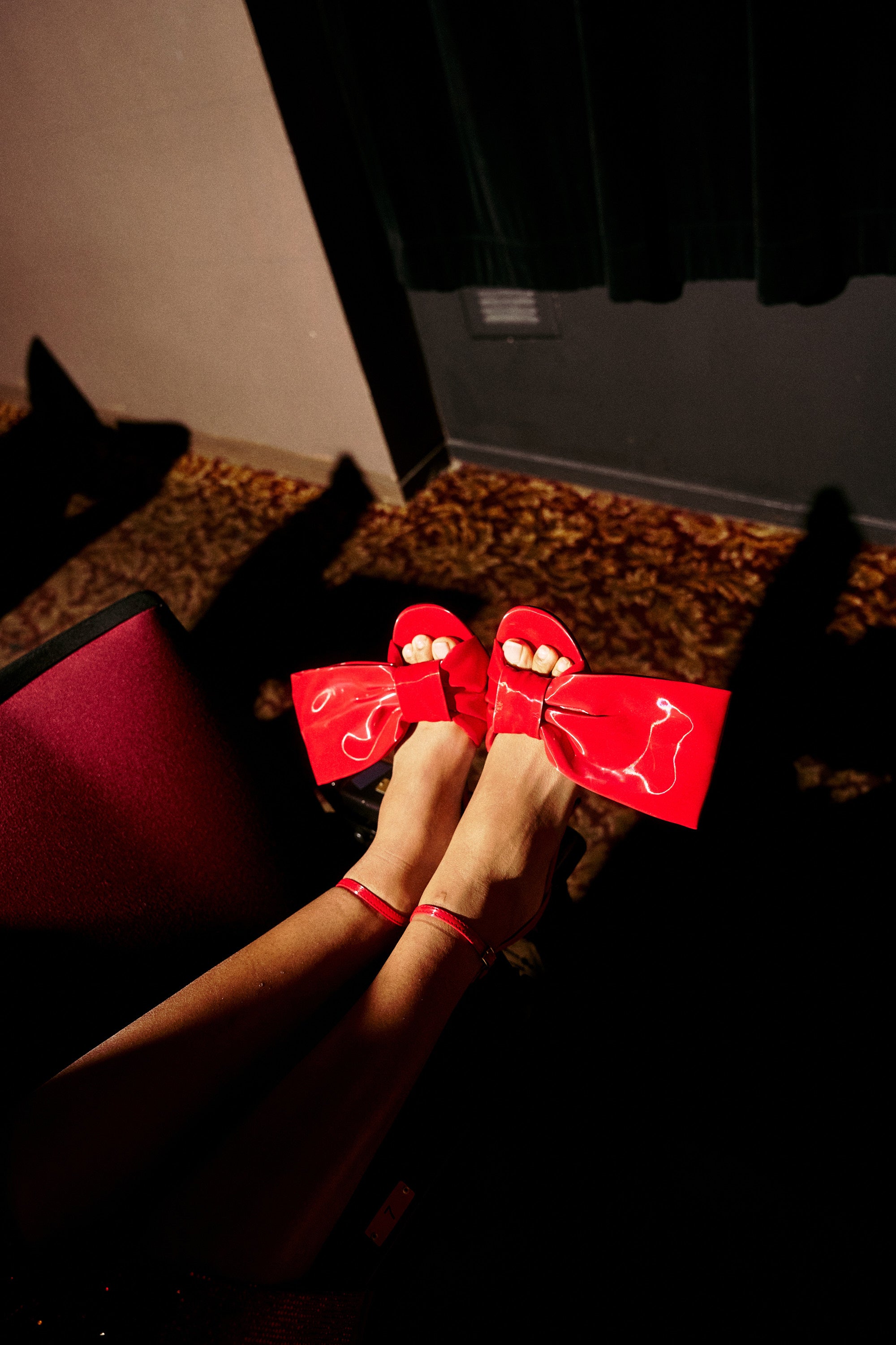 A person wearing AZALEA WANG LANYIN RED LIQUID LUXE BOW SANDALS sits with crossed legs in a dimly lit room, shadows falling across the patterned carpet and wall.