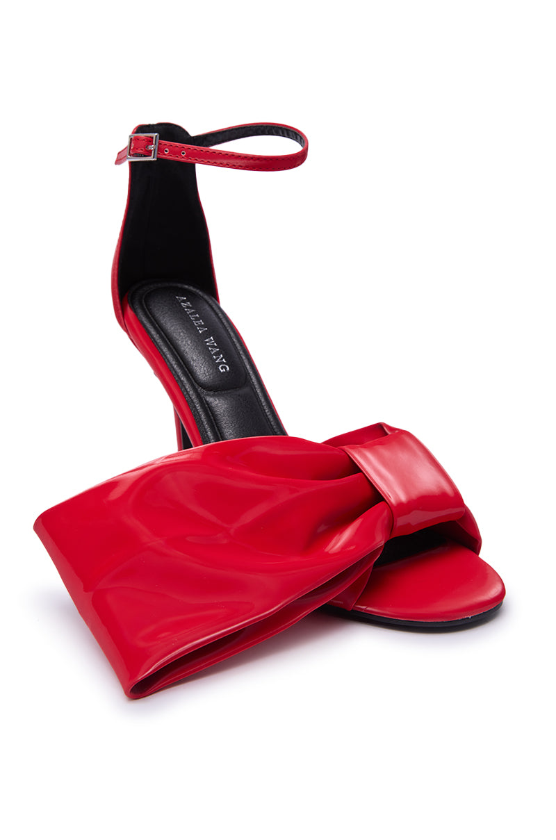 The AZALEA WANG LANYIN RED LIQUID LUXE BOW SANDAL is a faux leather high-heeled ankle strap sandal with a large shiny red bow, stiletto heel, and black “HALLA FANG” insole, set against a white background.