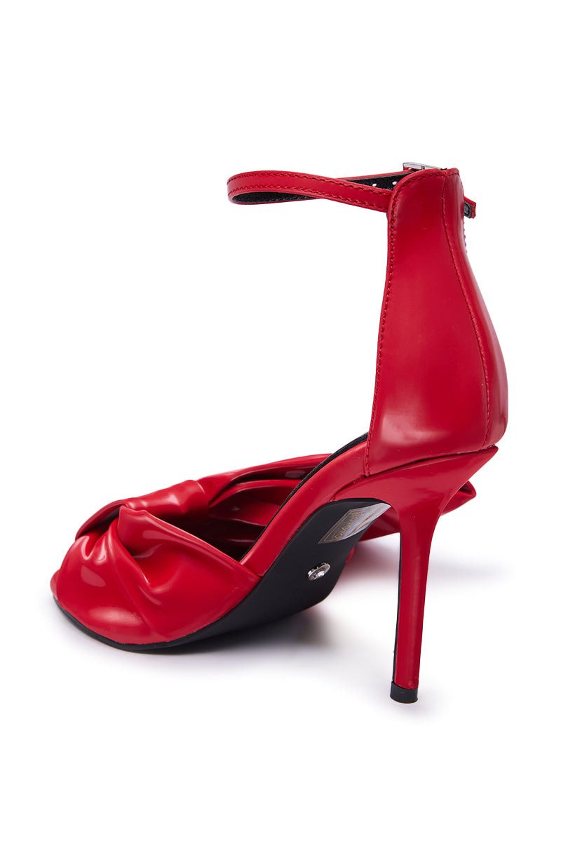 The AZALEA WANG LANYIN RED LIQUID LUXE BOW SANDAL is a shiny red faux leather high-heeled sandal with an ankle strap, peep toe, front bow detail, and a stiletto heel with a back zipper, shown angled for a side and rear view.