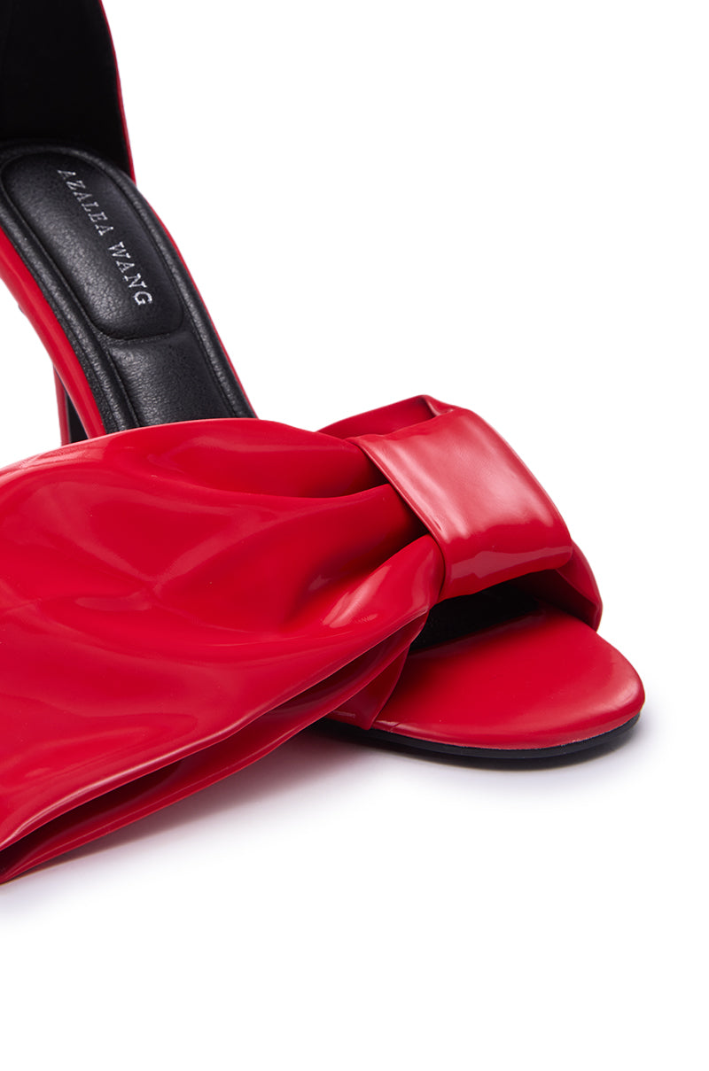 Close-up of the AZALEA WANG LANYIN RED LIQUID LUXE BOW SANDAL, featuring a glossy red faux leather bow across the toe, a stiletto heel, and a black insole with "LABELLE YANG," shown against a white background.