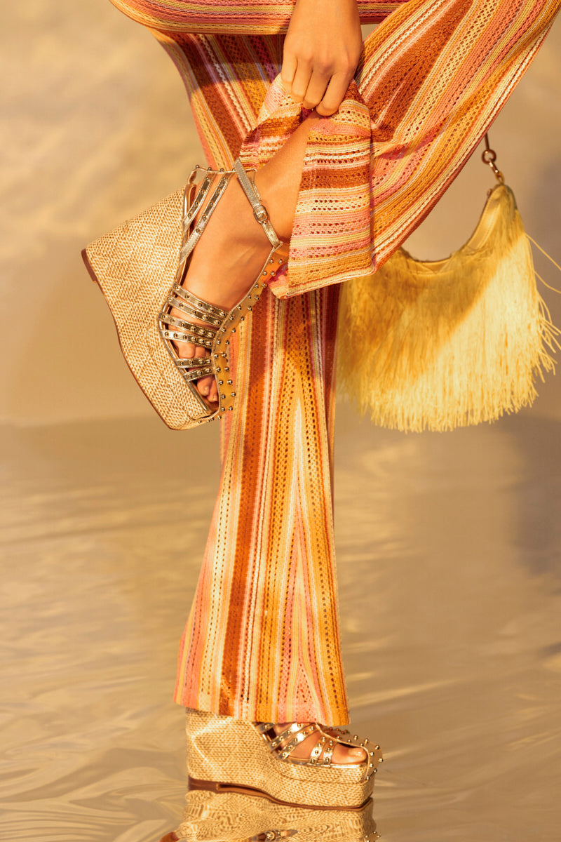 A woman confidently stands on a reflective surface, highlighting her AZALEA WANG LASZLO GOLD WEDGE SANDALS with metallic accents. Her fringed bag swings elegantly by her side, while warm tones of orange, pink, and gold complete the harmonious ensemble.