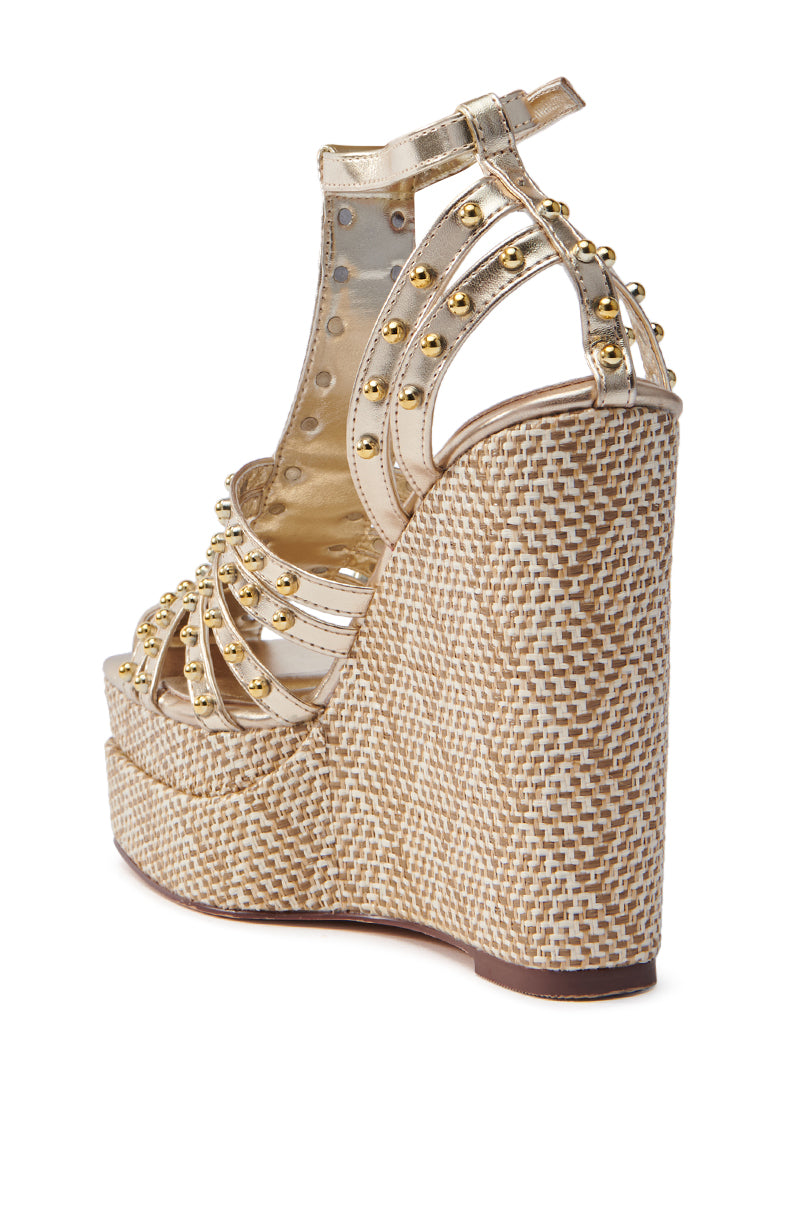 The AZALEA WANG LASZLO GOLD WEDGE SANDAL features a close-up of a shimmering metallic gold upper with studs and straps, an open-toe design, chic buckle ankle fastening, and a woven raffia platform sole and heel.