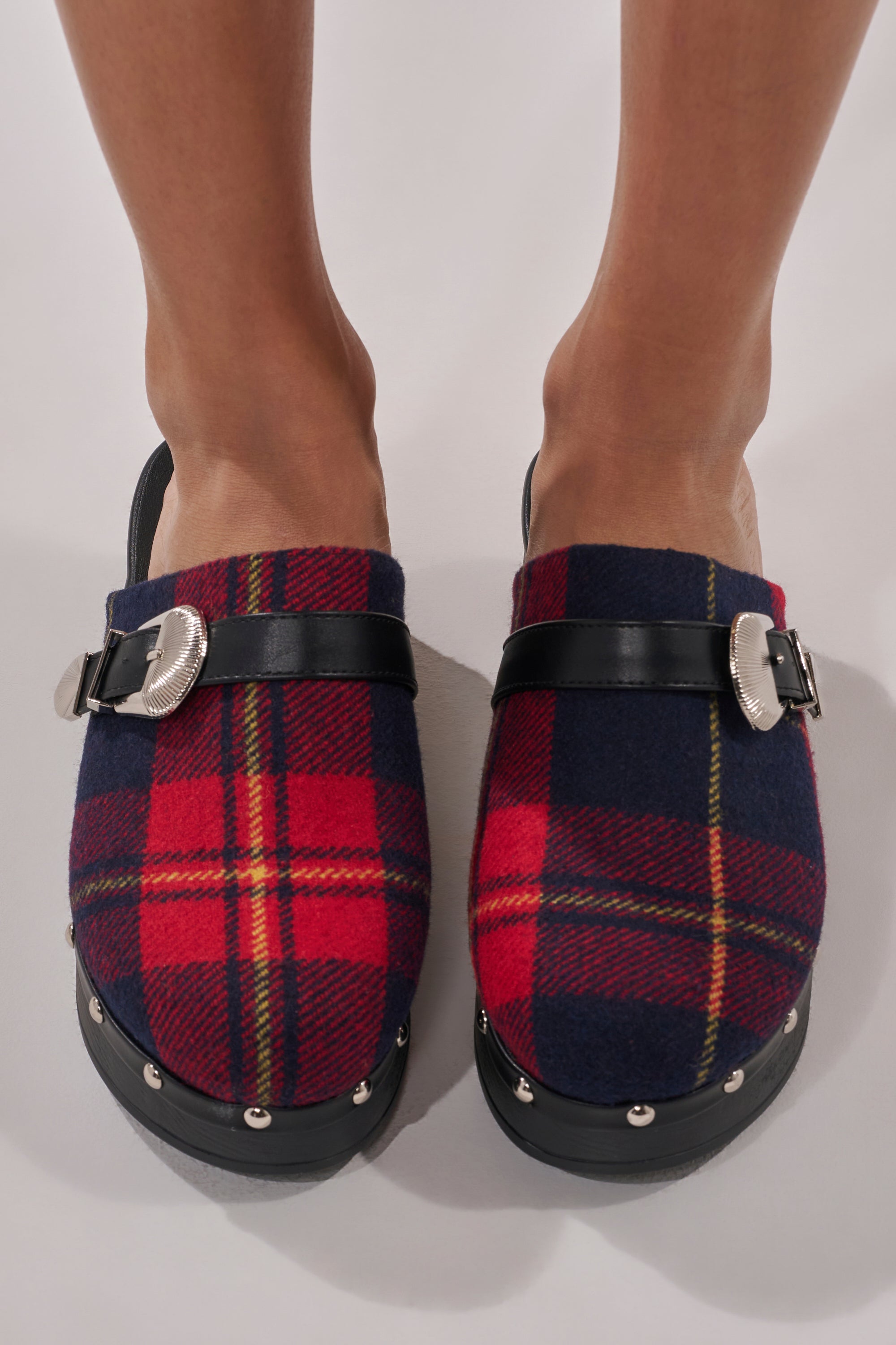 A person wearing AZALEA WANG LATIFAH RED PLAID HARDWARE FLAT MULE clogs with black straps, silver buckles, and silver-studded black soles stands on a light-colored surface.