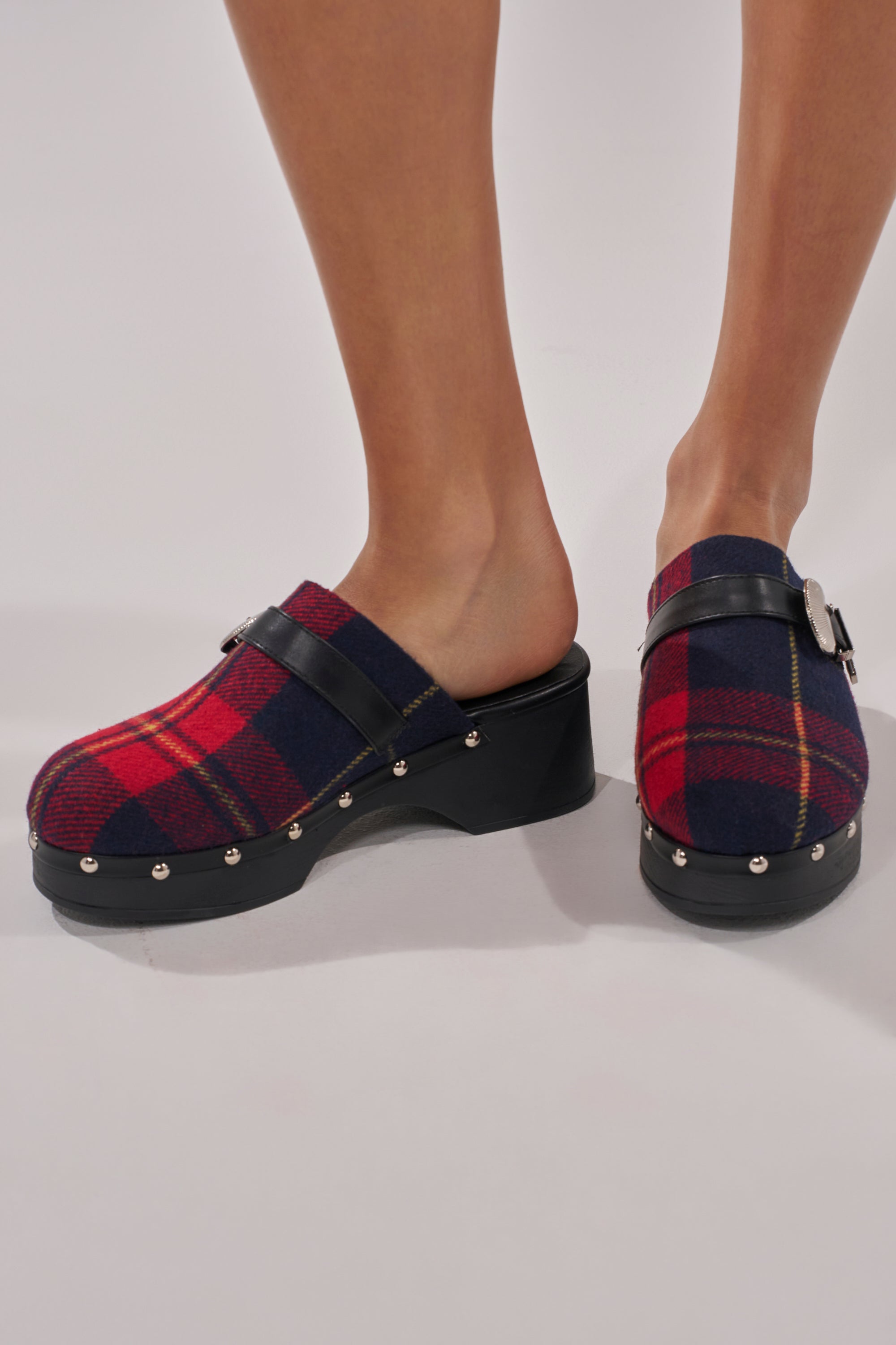 A person wearing the AZALEA WANG LATIFAH RED PLAID HARDWARE FLAT MULE, featuring red and navy plaid fabric, black leather straps with silver buckles, cushioned insole, and silver studs on black soles, stands against a white background.