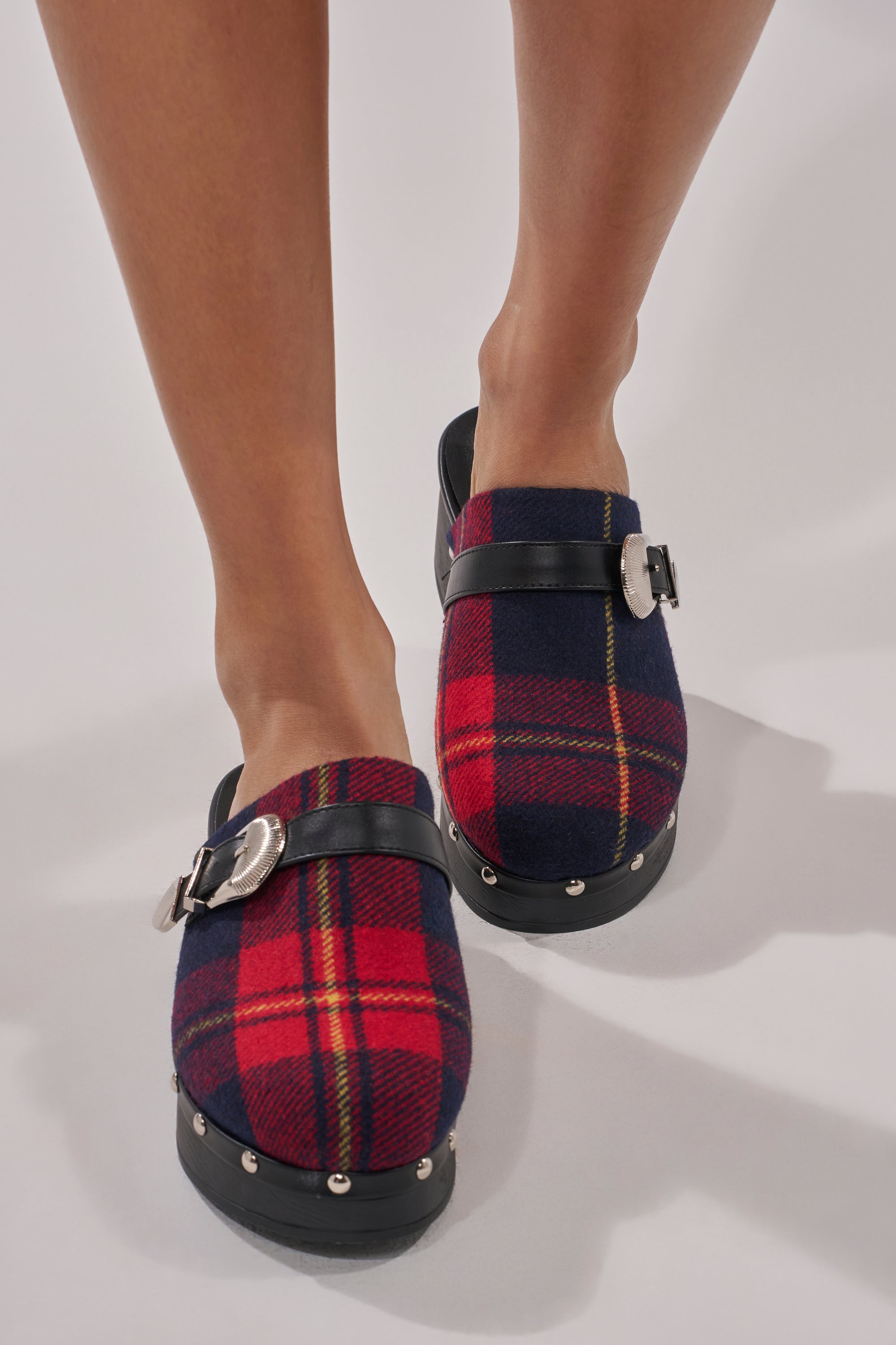 A person wears the AZALEA WANG LATIFAH RED PLAID HARDWARE FLAT MULE, featuring red and navy plaid fabric, black straps with silver buckles, studded soles, and cushioned insoles, standing on a light surface.
