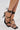 A close-up of a person wearing the AZALEA WANG LAURA BLACK STILETTO SANDAL, featuring black strappy design, square toe heel, and thin lace-up fastening wrapping around the ankles; toes have a light polish.