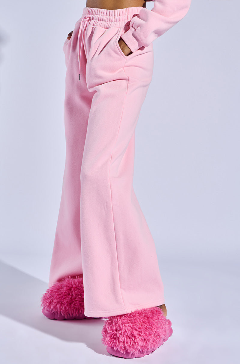 A person wearing AZALEA WANG LAWSON PINK SLIPPER with faux fur upper and light pink sweatpants stands hands in pockets. The plain light background highlights the cozy footwear.
