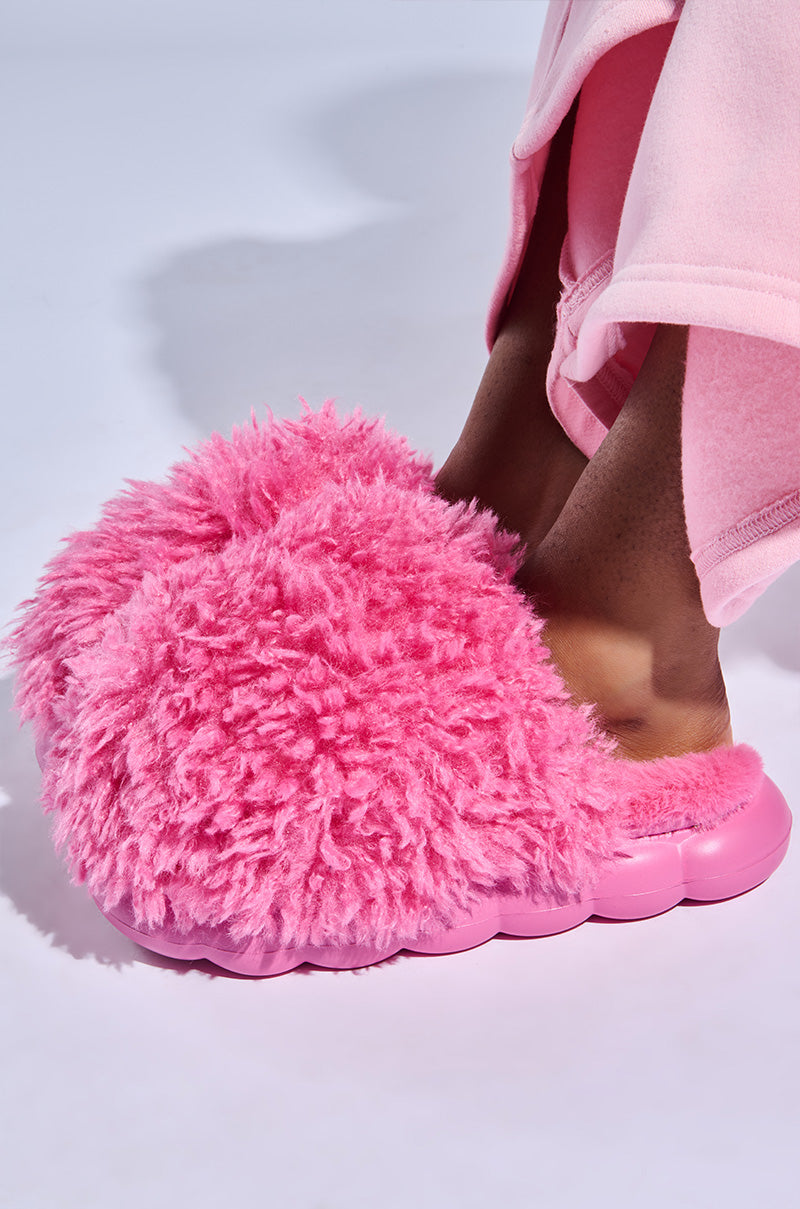 A close-up shows a person in pink attire wearing the AZALEA WANG LAWSON PINK SLIPPER, featuring large, fluffy faux fur uppers and cloud-like flatform soles, set against a light gray background.