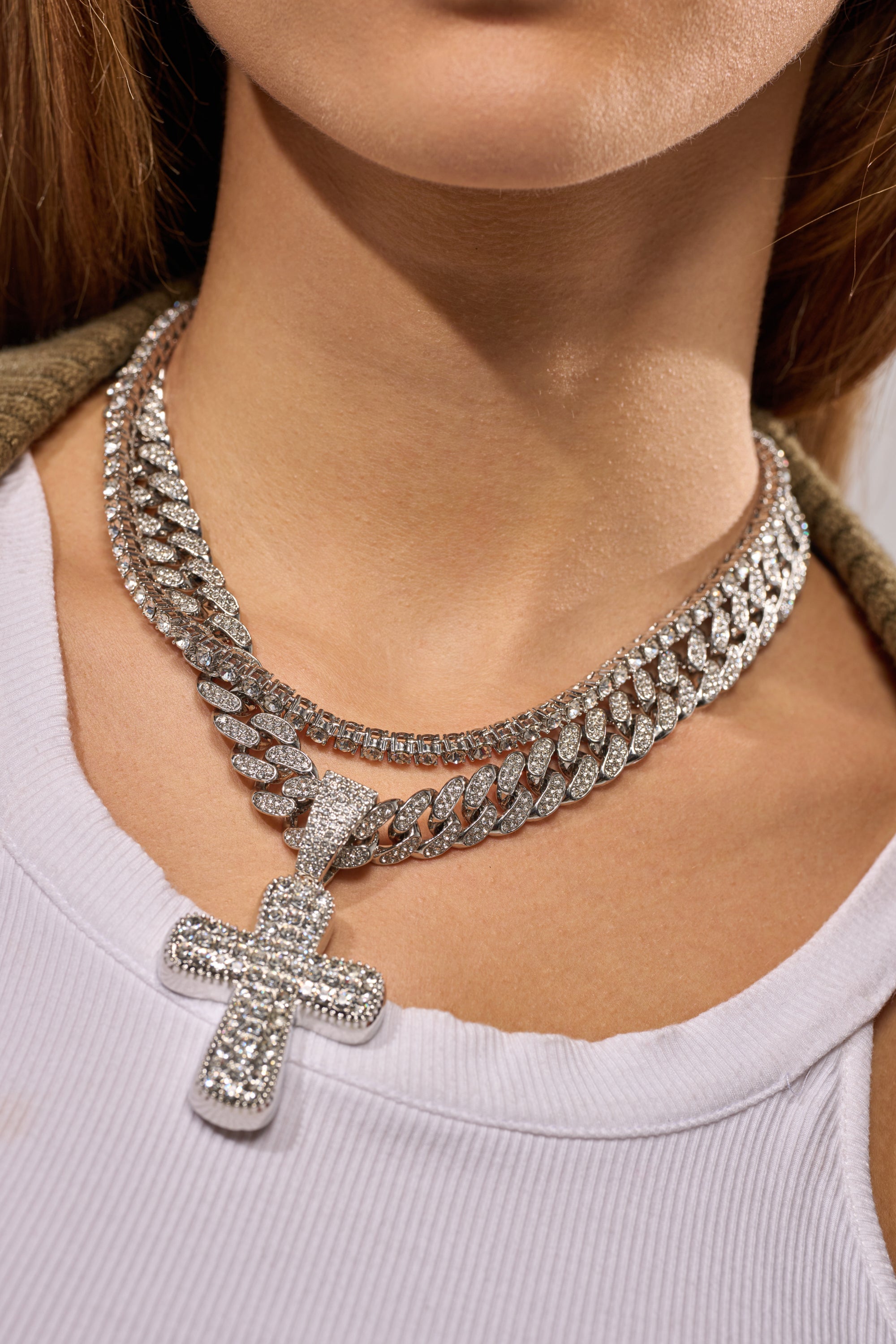 A woman wears the GIMME NECKLACE, a layered set featuring sparkling rhinestones, a bold Cuban link chain, and a large rhinestone cross pendant, styled over a white ribbed tank top. 3