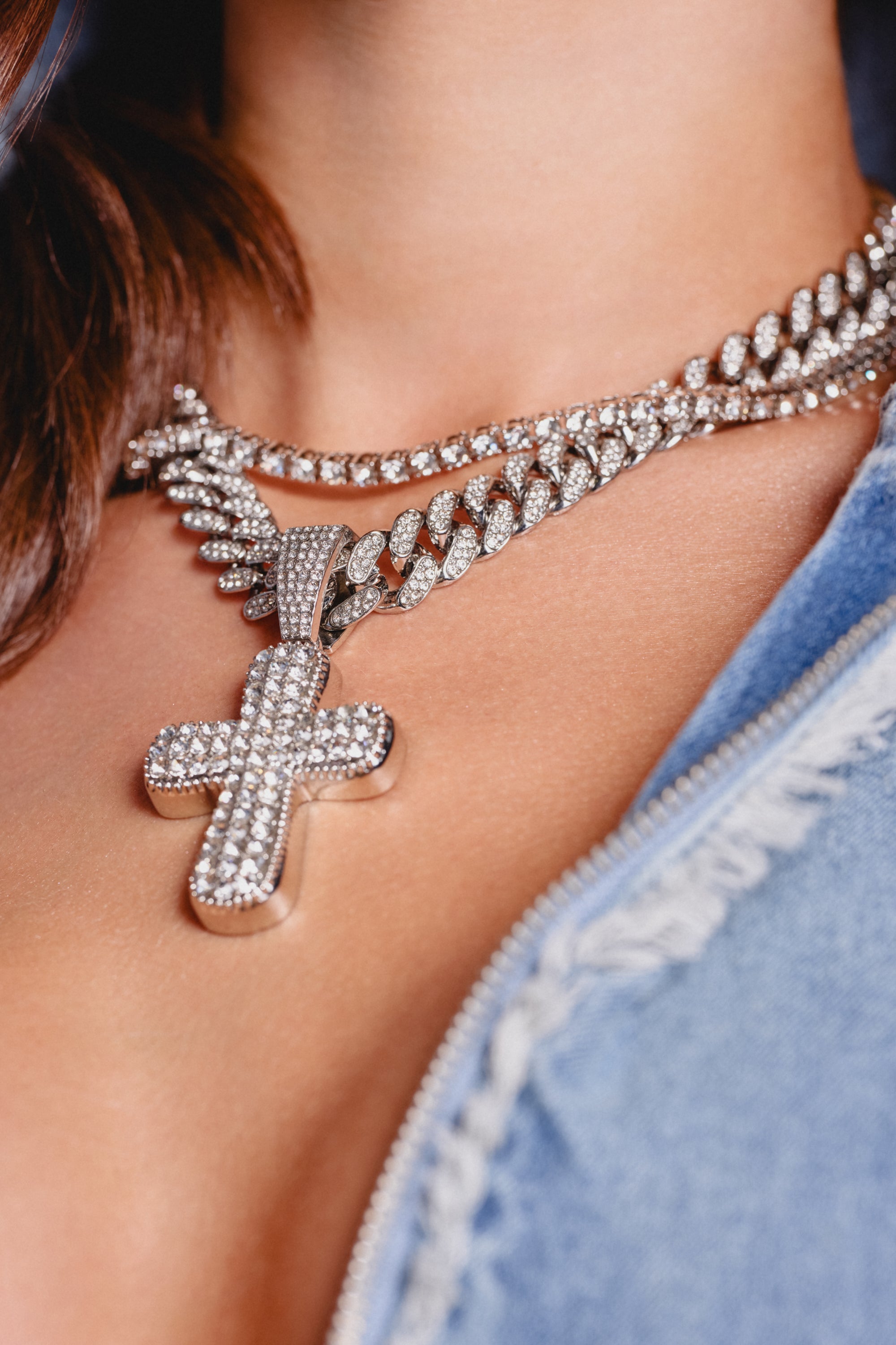 A close-up of a person wearing the GIMME NECKLACE, a layered set with two sparkling diamond chains and a rhinestone cross pendant, styled over bare skin and a partially unzipped blue denim jacket. 1