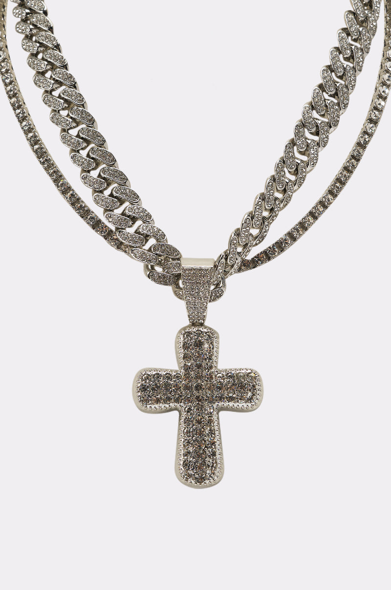 The GIMME NECKLACE is a stunning layered set featuring three silver chains with diamond-like stones and a bold rhinestone cross pendant, beautifully displayed against a plain white background. 6