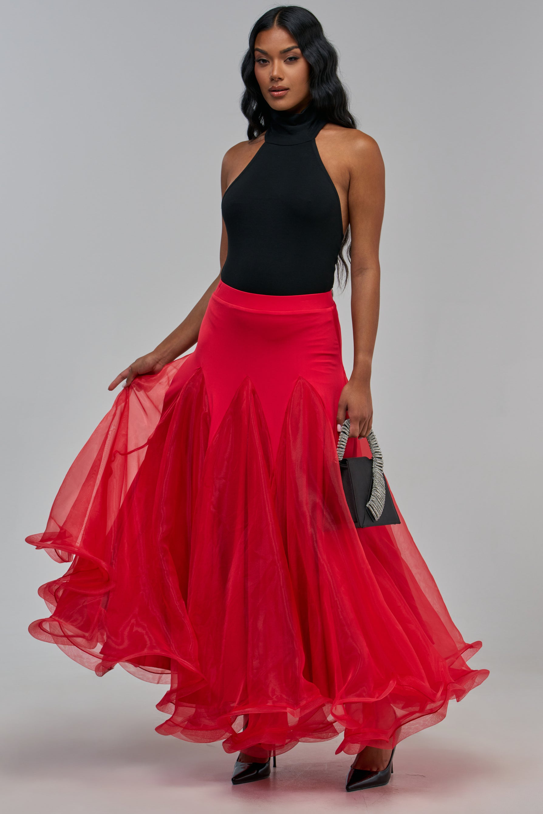 A model wears a black halter top and the RED HOT RUFFLE SKIRT, a red mesh maxi skirt with a flared A-line silhouette, while holding a small black handbag.