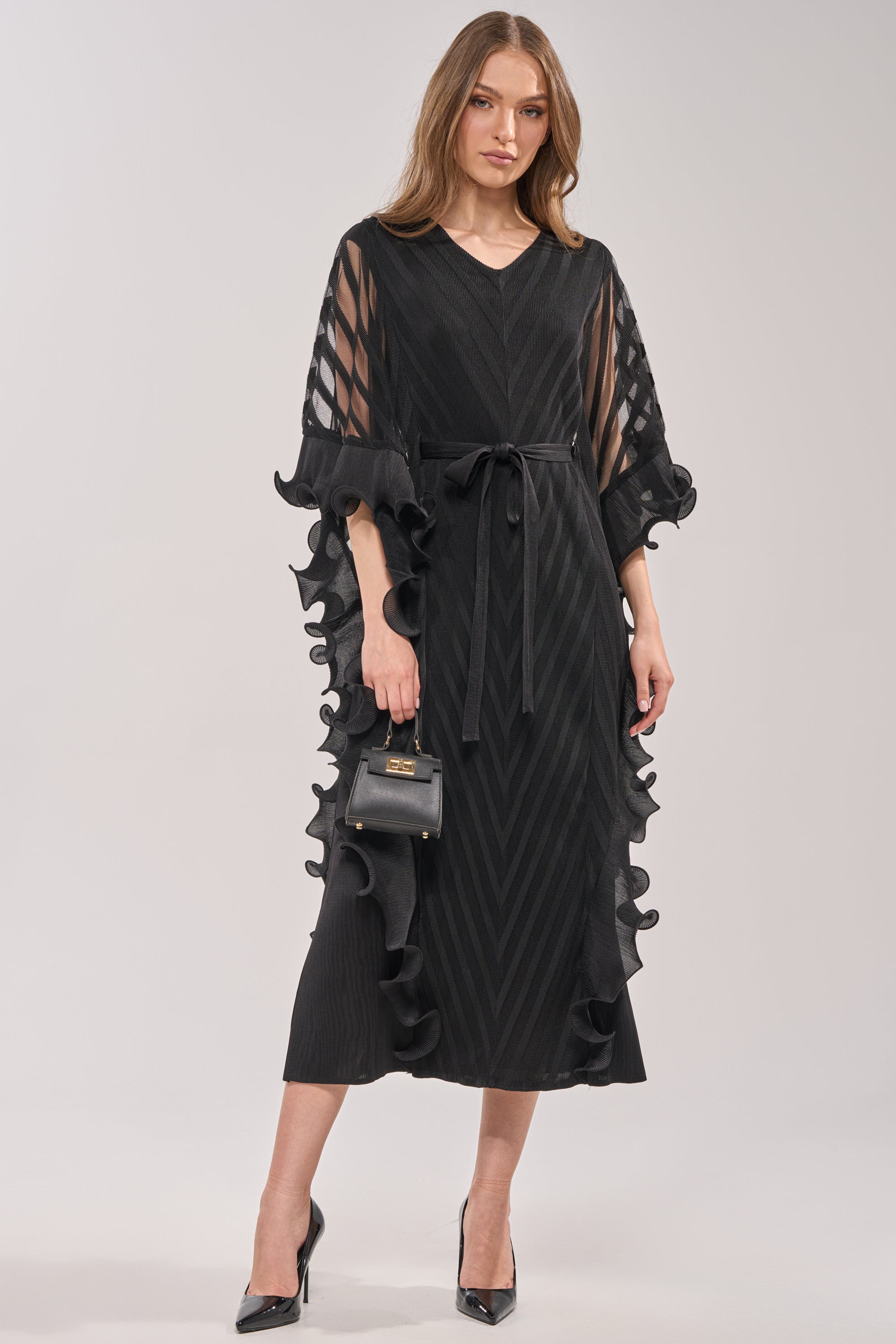 A woman faces forward in THE NIGHT GOES ON RUFFLE MAXI DRESS IN BLACK, featuring sheer striped sleeves, ruffled edges, a belted waist, and black high heels. She holds a small black handbag and wears her hair loose—ideal for formal occasions. 0