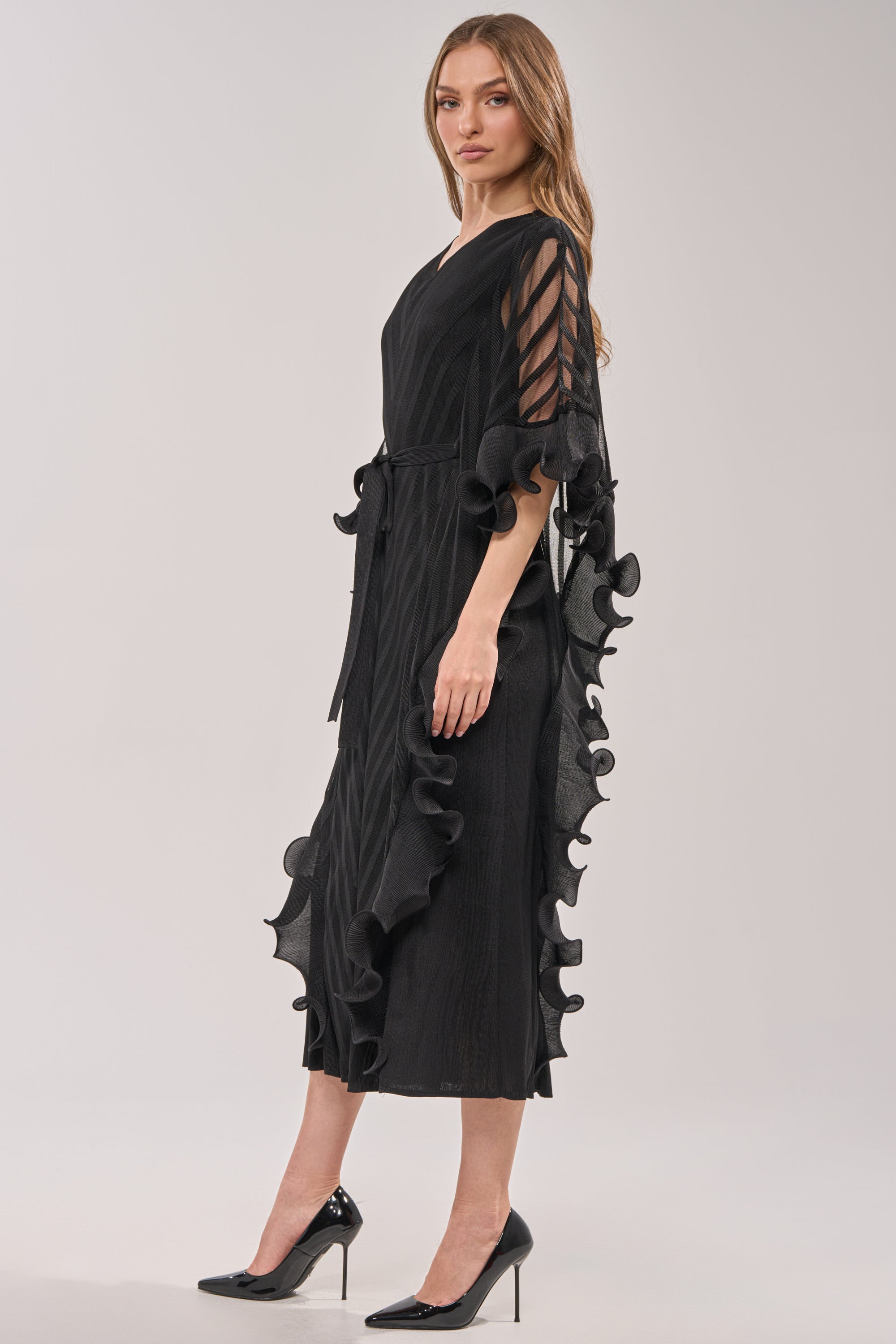 A woman models THE NIGHT GOES ON RUFFLE MAXI DRESS IN BLACK, featuring ruffle overlays and sheer striped sleeves, standing in high heels against a plain light background with a neutral expression. 2