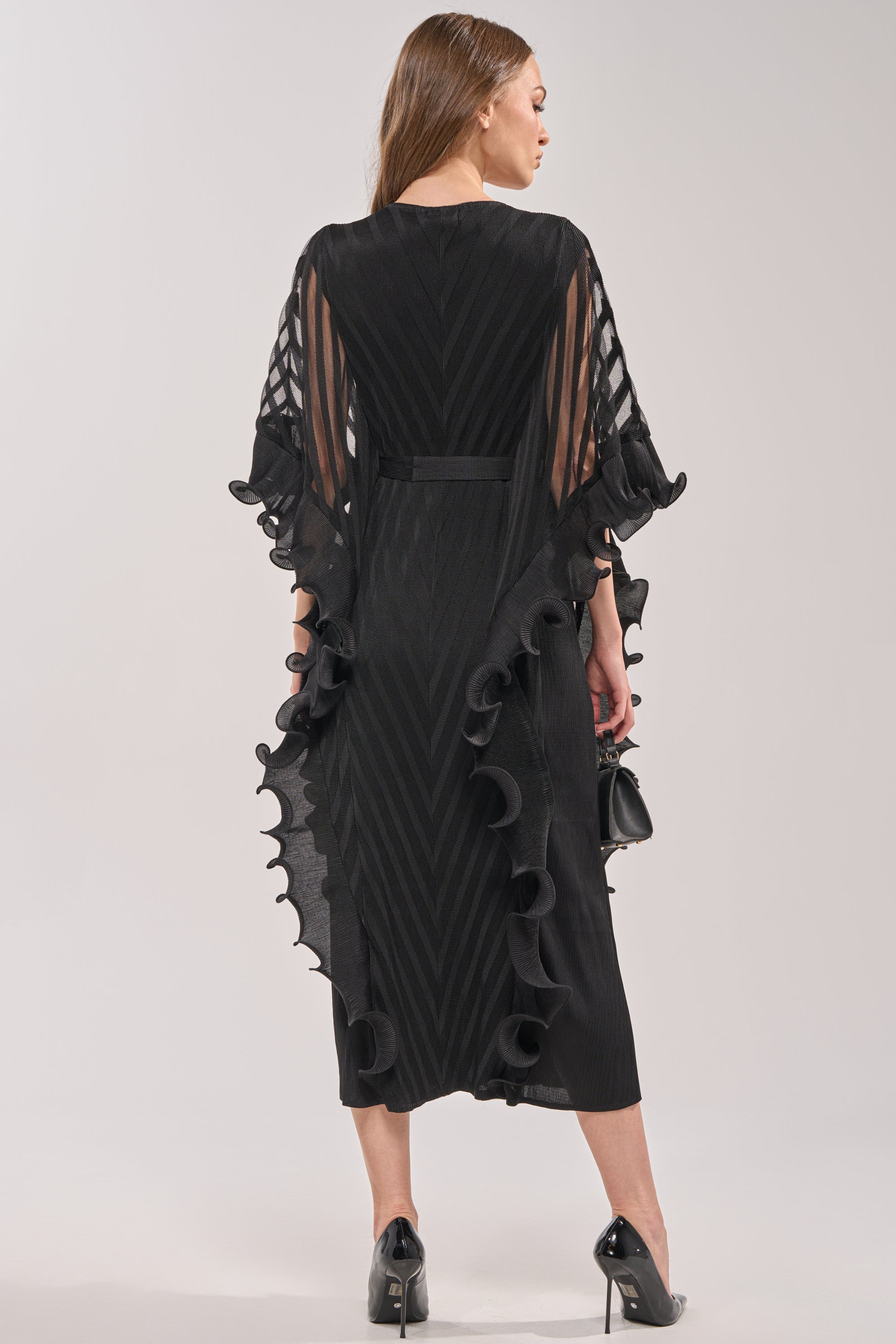 A woman faces away, wearing THE NIGHT GOES ON RUFFLE MAXI DRESS IN BLACK with sheer long sleeves and chevron fabric, paired with a small black purse and black high heels. 3