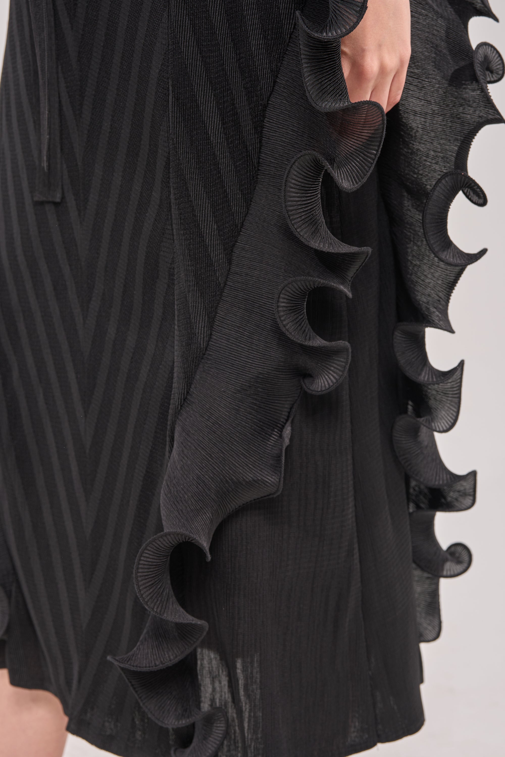 Close-up of THE NIGHT GOES ON RUFFLE MAXI DRESS IN BLACK, showcasing its chevron stripe pattern and decorative wavy ruffles along the side. A hand is gently placed near the ruffles. 4