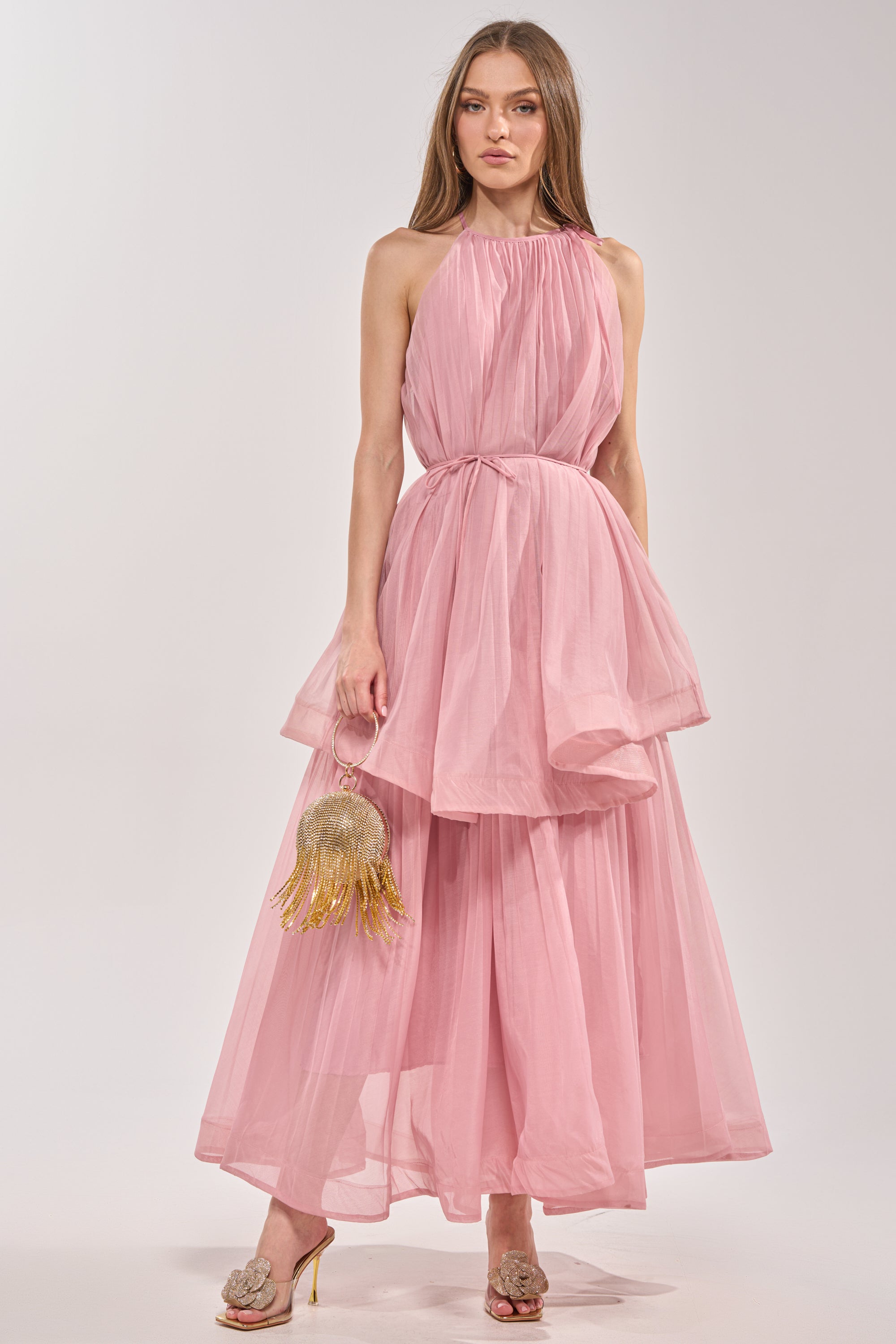 A woman wears the BREEZY GIRL TIERED MAXI DRESS IN BLUSH, a flowing maxi with pleated chiffon and ruffle tiers. She holds a gold fringe handbag, pairs heeled open-toe shoes, and lets her long hair down against a plain, light background. 3