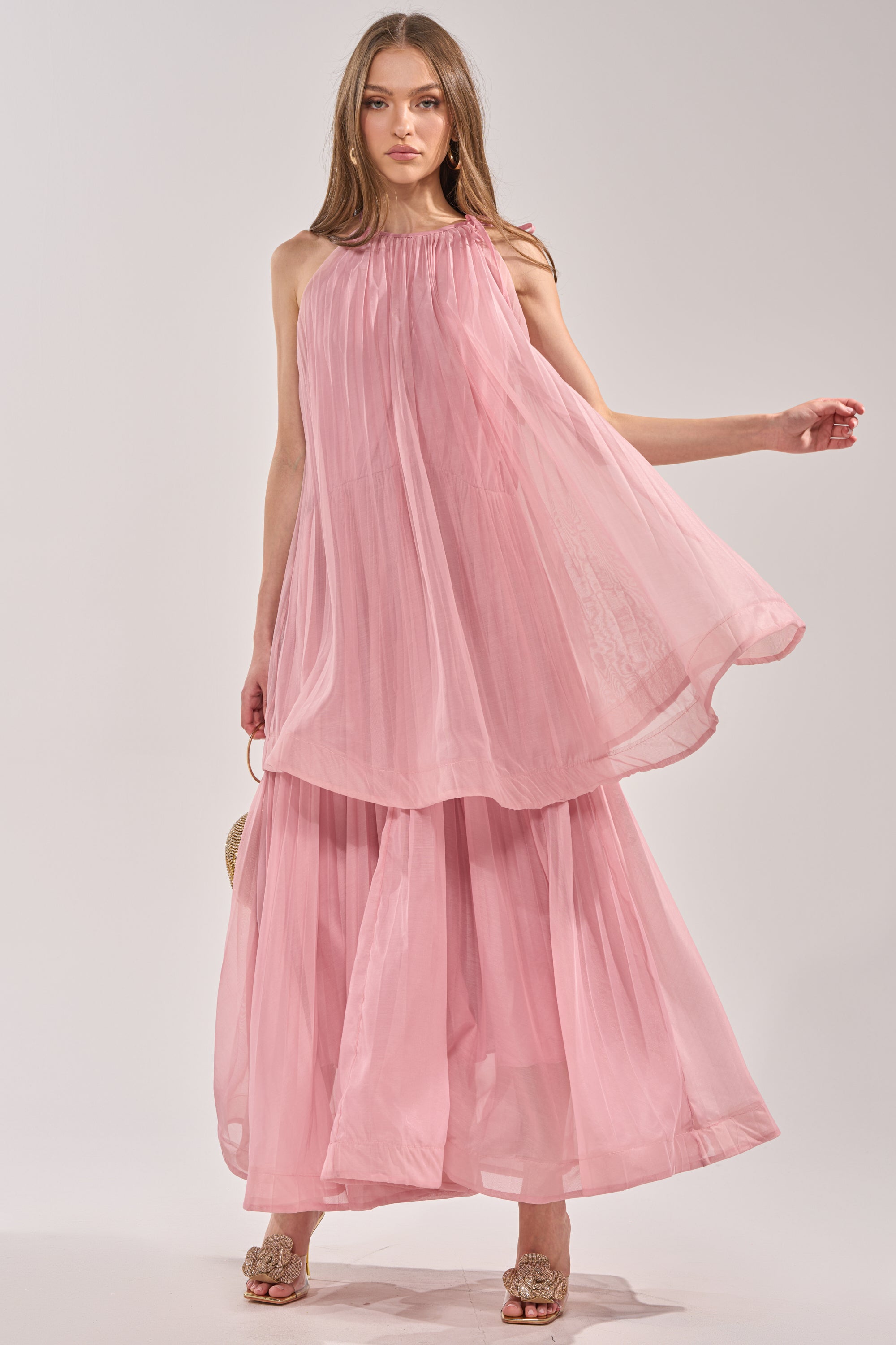 A woman models the BREEZY GIRL TIERED MAXI DRESS IN BLUSH, a sleeveless blush pink maxi with a billowy layered top, tiered ruffle details, and a wide skirt. She pairs it with beige sandals against a plain light gray background. 0