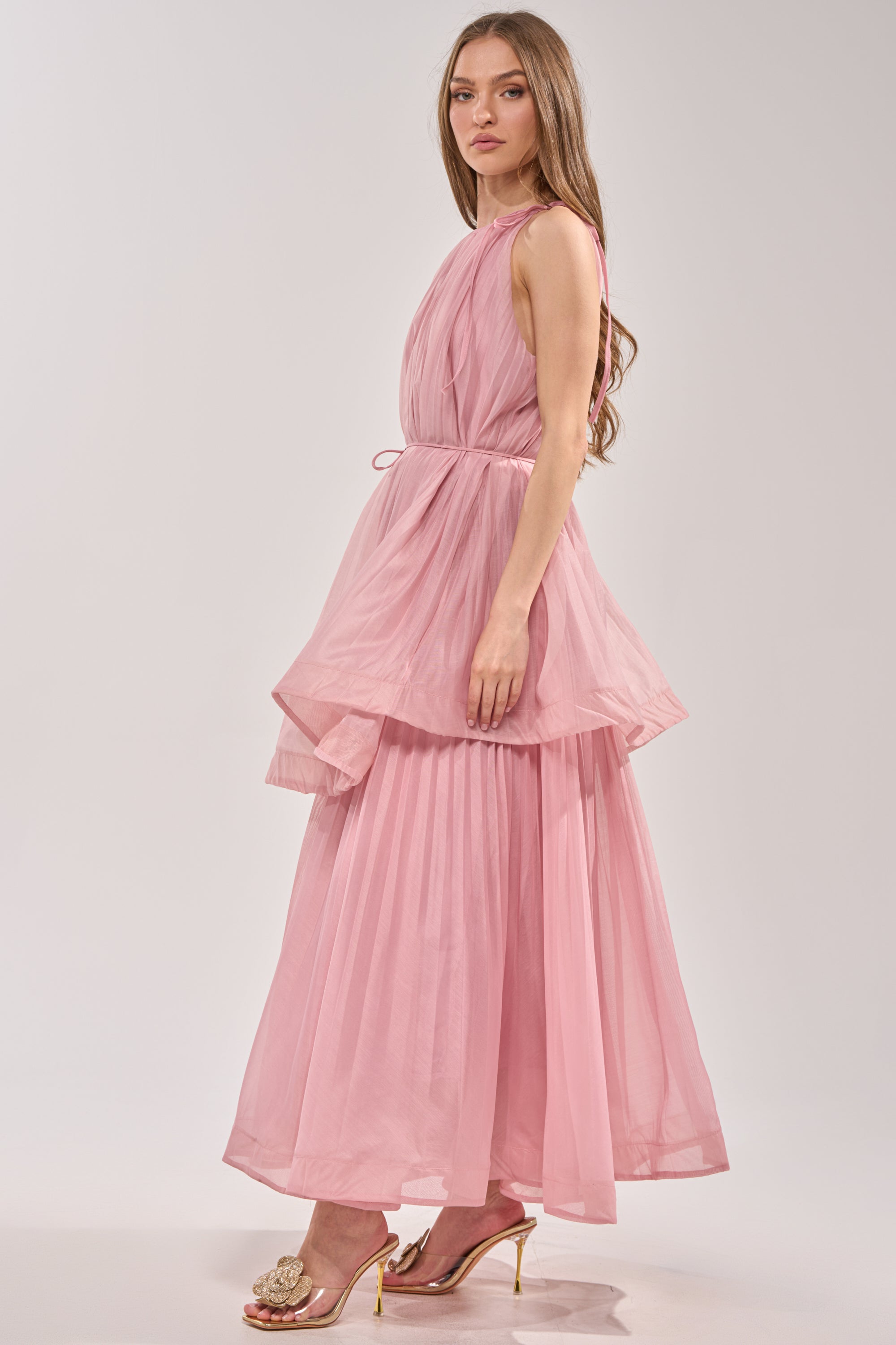 A woman in profile wears the BREEZY GIRL TIERED MAXI DRESS IN BLUSH— a sleeveless pleated chiffon maxi with ruffle tiers and a thin waist tie, styled with long straight hair and flower-embellished open-toe heels against a plain white background. 5