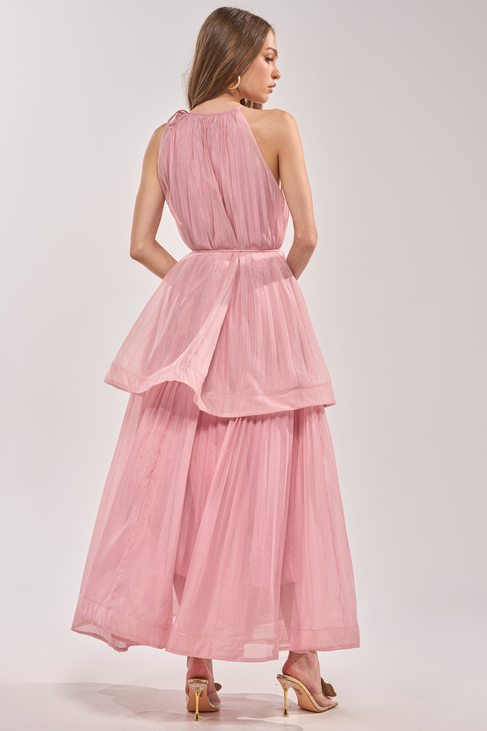 A woman faces away in the BREEZY GIRL TIERED MAXI DRESS IN BLUSH, featuring pleated chiffon and a tiered ruffle skirt, paired with gold heels against a plain light background. 6