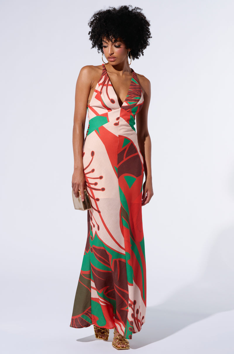 A woman with curly hair is wearing a vibrant HIBISCUS HONEY PRINTED MAXI DRESS featuring an abstract geometric patterning in red, green, and brown. She holds a small beige clutch in her right hand and stands against a plain white background, accessorizing her ensemble with gold platform sandals.