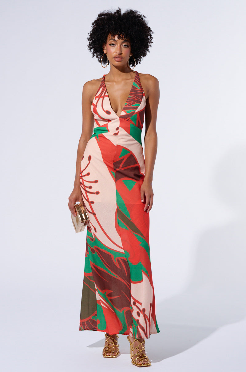A woman with curly hair is wearing a sleeveless Hibiscus Honey Printed Maxi Dress with a plunging neckline and an abstract geometric pattern in red, green, and beige. She holds a small beige clutch and sports gold platform sandals while standing against a plain white background.