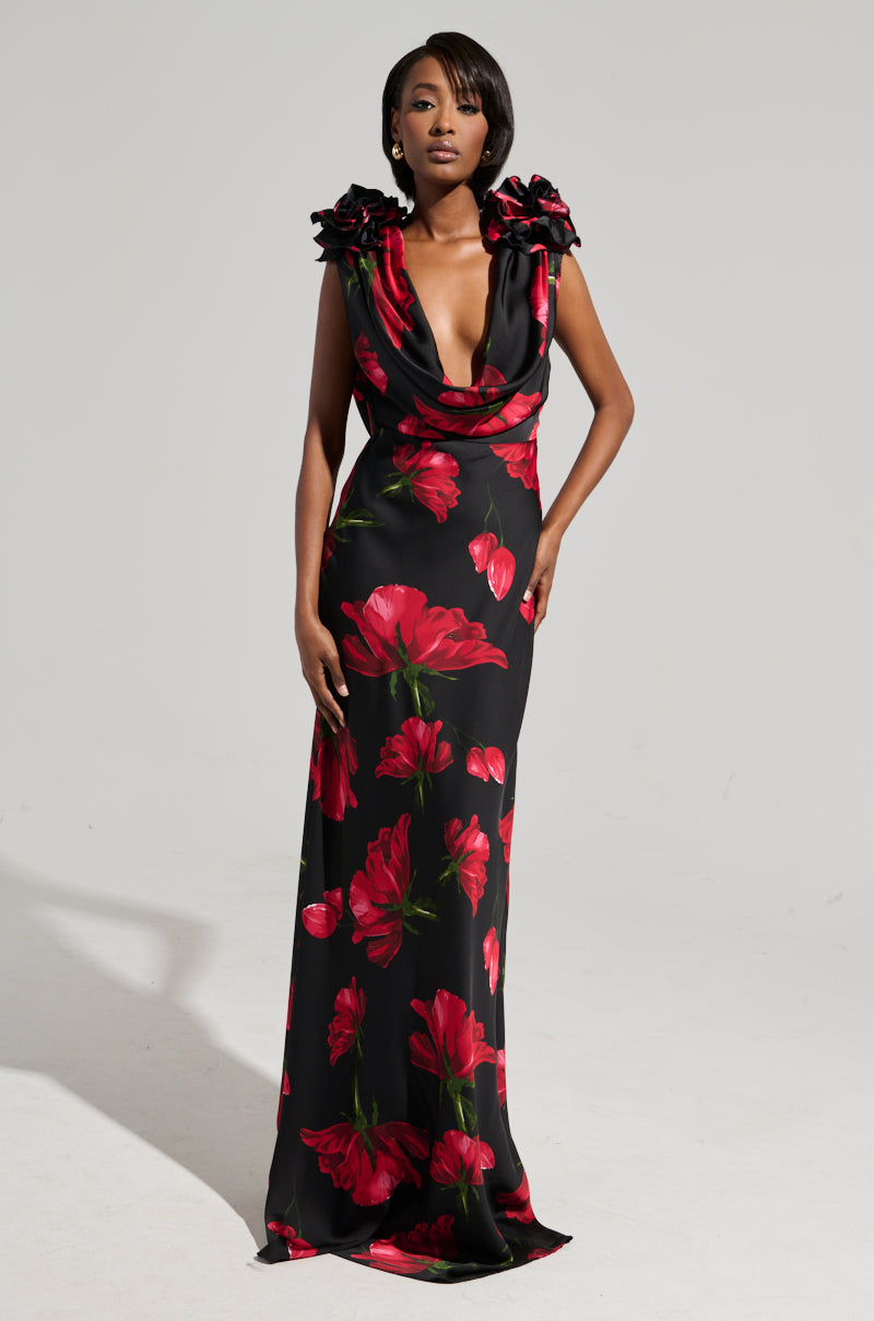 A person stands confidently wearing the ROSE MAXI DRESS, a long and elegant black dress adorned with large red floral patterns. Crafted from satin, this dress features a deep neckline and decorative shoulder details. The background is plain and minimalist.