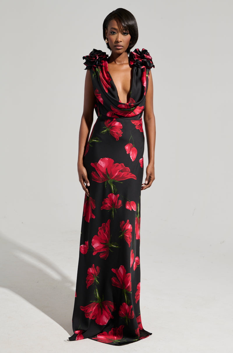 A woman is set against a simple backdrop, showcasing the eye-catching ROSE MAXI DRESS. This maxi dress flaunts a long, sleeveless design embellished with bold red floral patterns. It features an elegant satin finish with a deep neckline and floral details on the shoulders. Her hair is styled in a chic, short look.